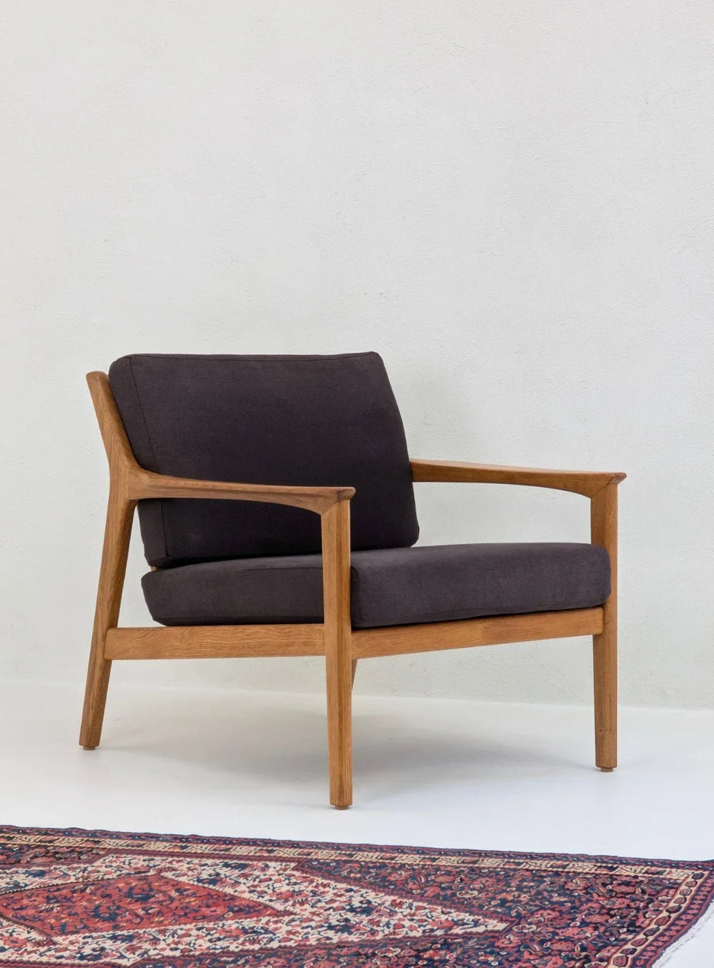 Mid-century modern wooden armchair with dark cushions on a patterned rug in a minimal room