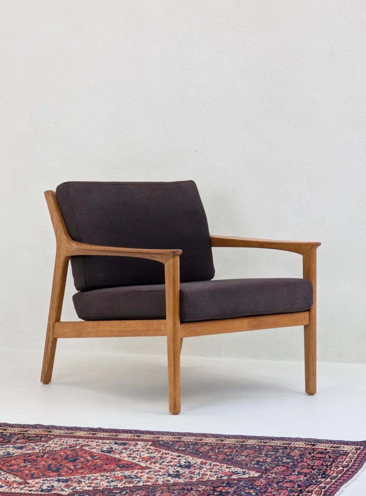 Mid-century modern wooden armchair with dark cushions on a patterned rug in a minimal room