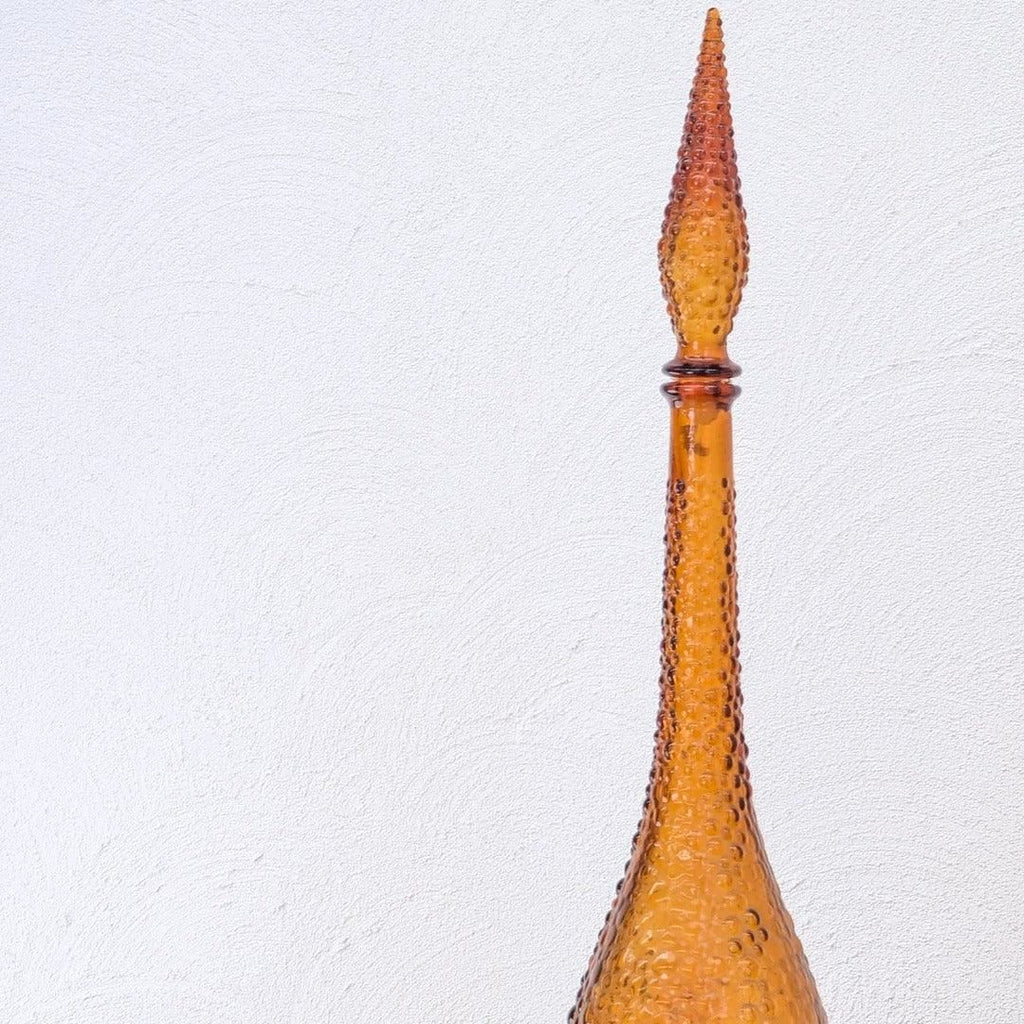 Tall amber hobnail glass decanter against a white textured wall