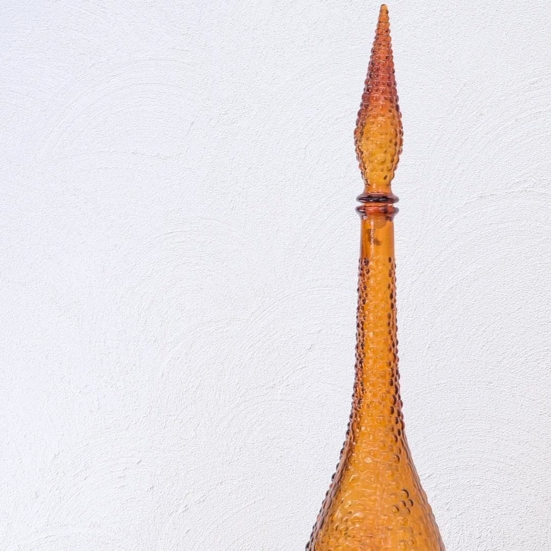 Tall amber hobnail glass decanter against a white textured wall