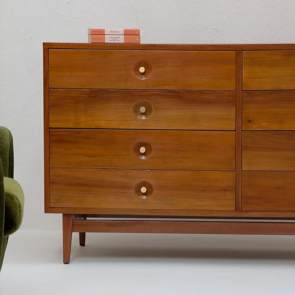 Mid-century modern wooden dresser with four drawers and brass knobs, books on top, green chair partial view