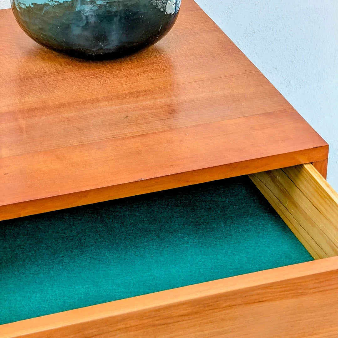 Open wooden drawer with teal felt lining seen under a cherry wood table, partial ceramic vase visible