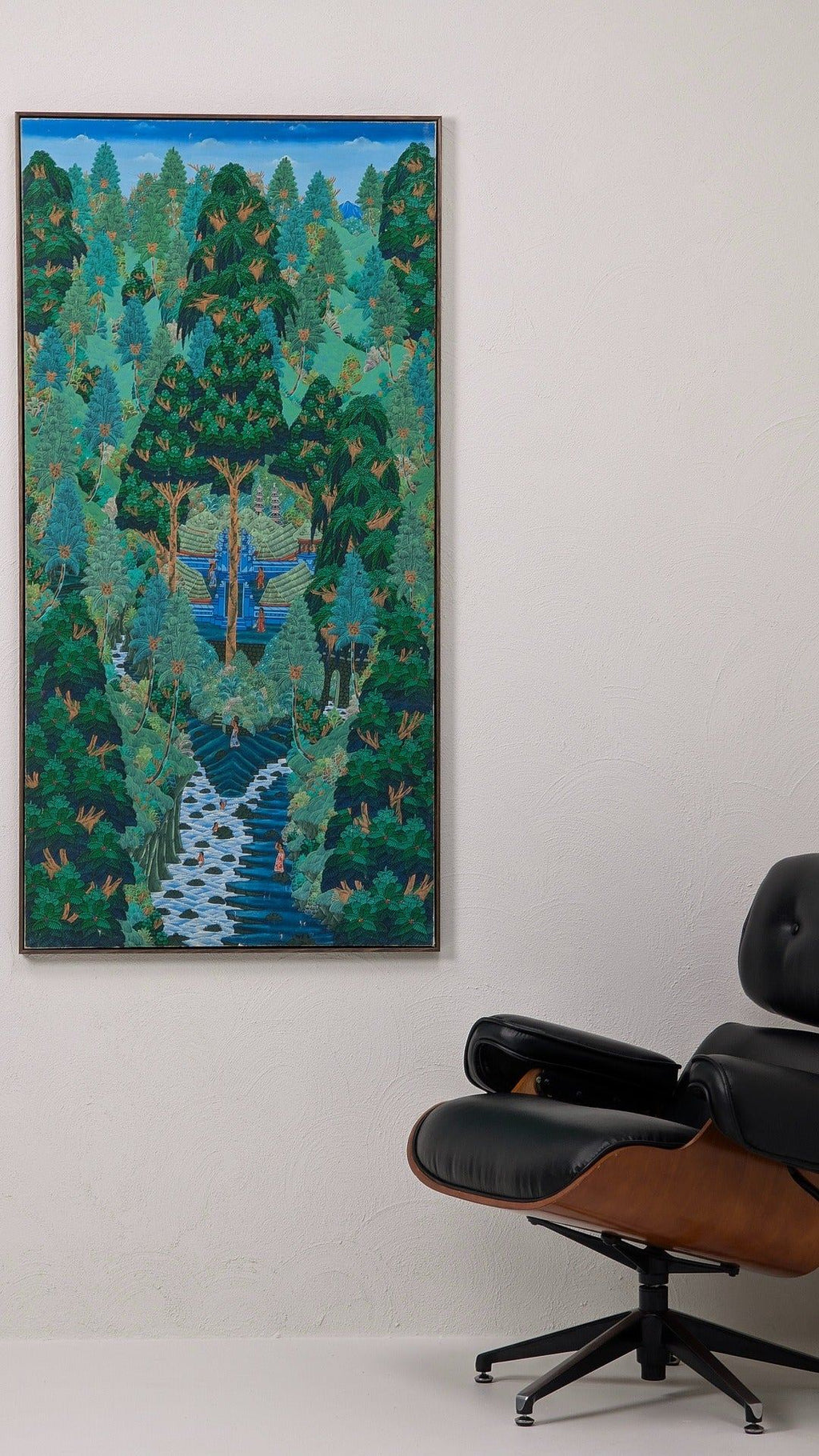 Framed colorful painting of lush forest and river scene with small figures, hanging on white wall above black leather lounge chair