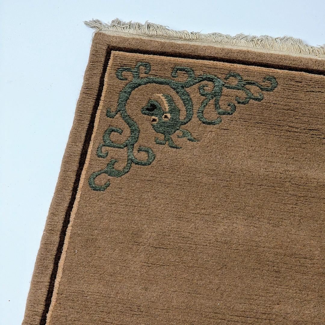 Corner of a brown wool rug with green ornate pattern and fringe on white background