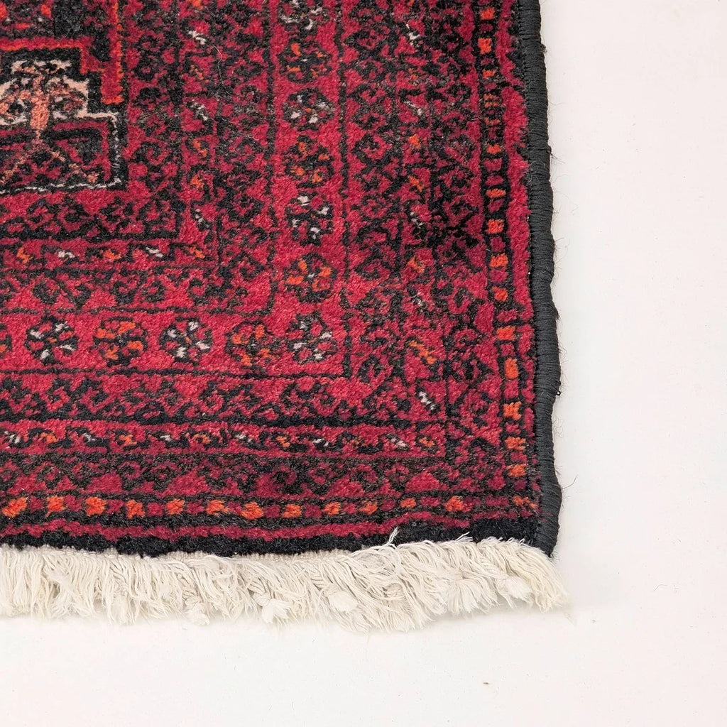 Corner detail of a red Persian rug with black patterns and white fringe on a light floor