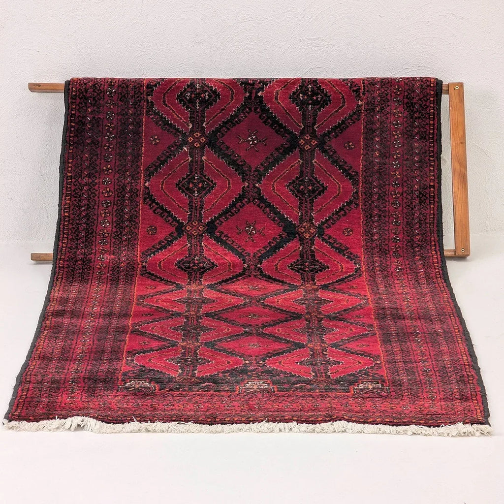 Red Persian rug with geometric black patterns on wooden display stand against white wall