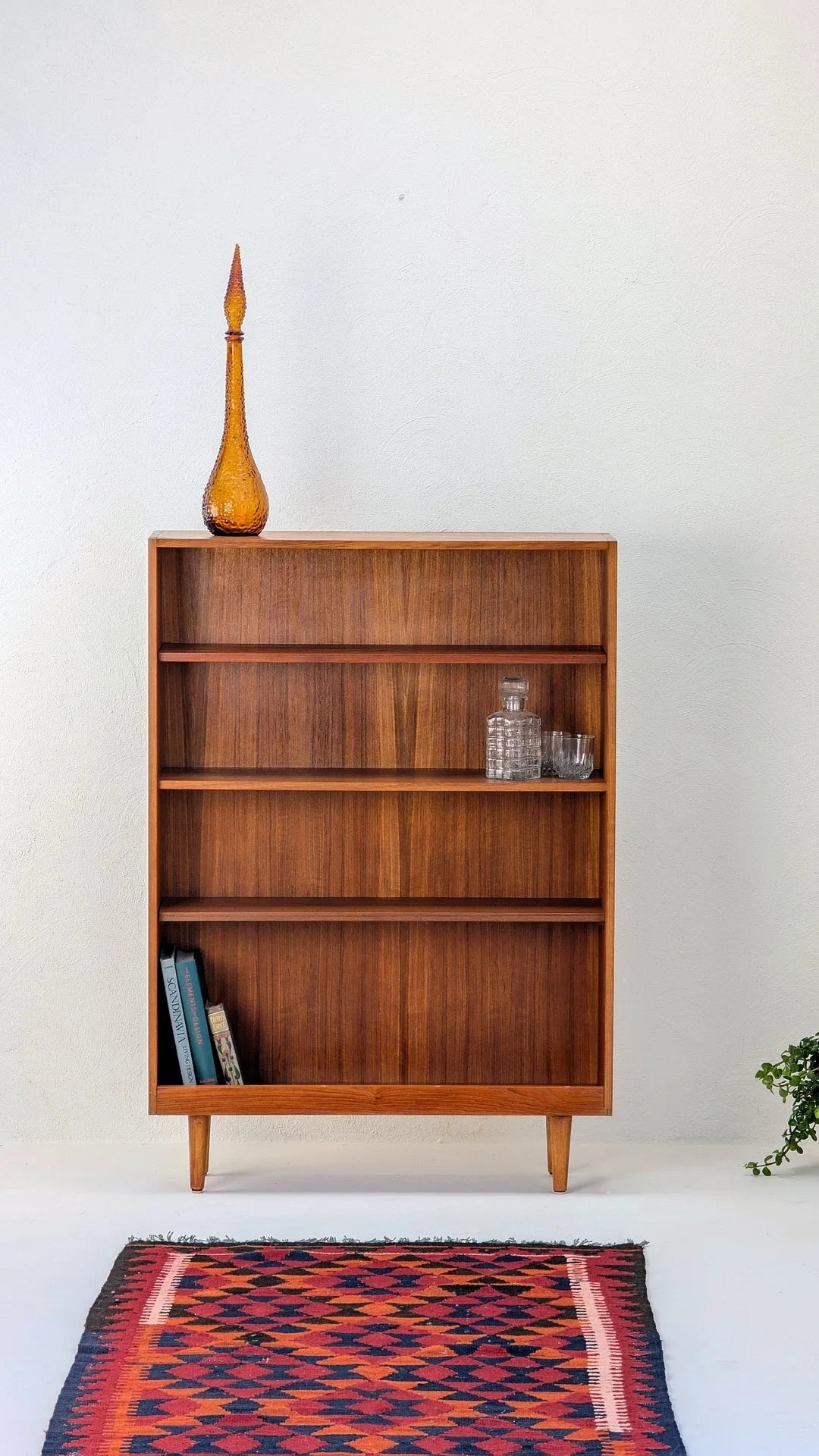 Mid-century wooden bookshelf with amber glass bottle, crystal decanter, books, and colorful patterned rug