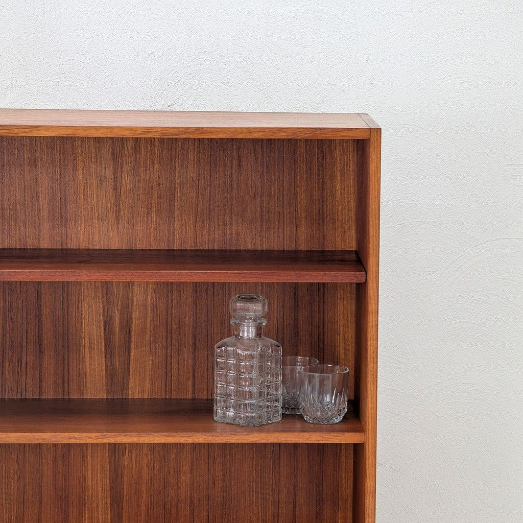Wooden bookshelf with a glass decanter and two clear glasses on middle shelf against white textured wall