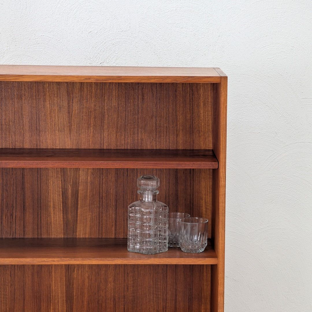Wooden bookshelf with a glass decanter and two clear glasses on middle shelf against white textured wall
