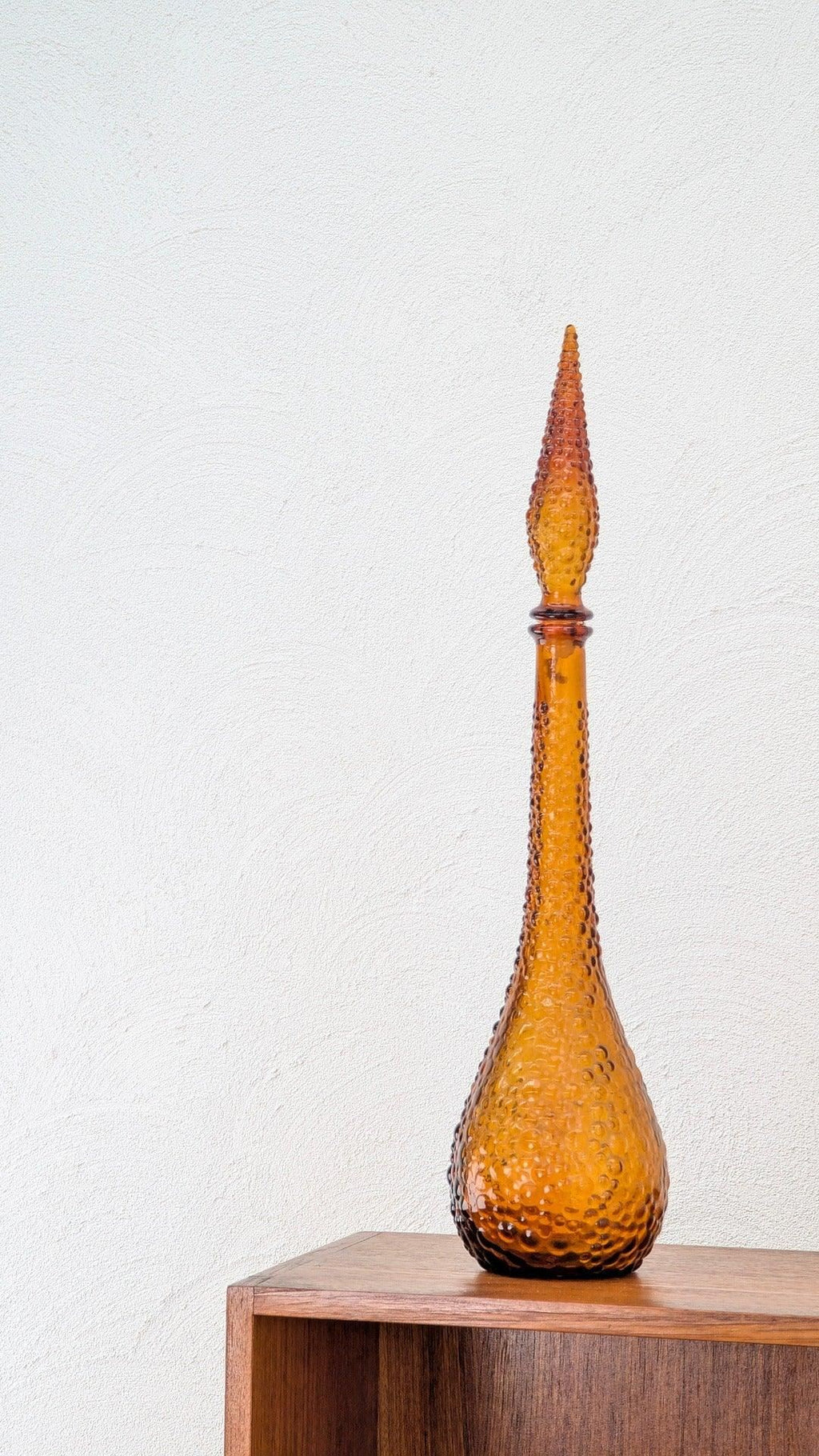 Amber hobnail glass decanter with pointed stopper on a wooden shelf against white wall