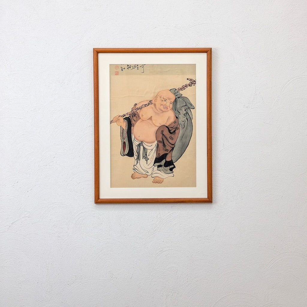 Framed traditional Asian painting of a smiling, bald monk holding a staff on textured white wall