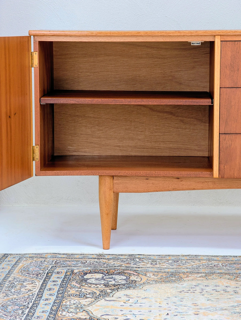 Mid-century wooden cabinet with open door revealing empty shelf, standing on tapered legs over patterned rug