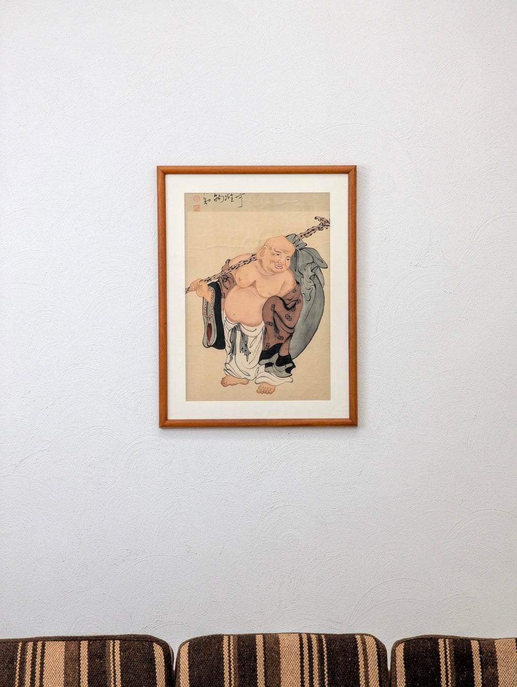 Framed Asian art of laughing Buddha on white wall above striped brown couch
