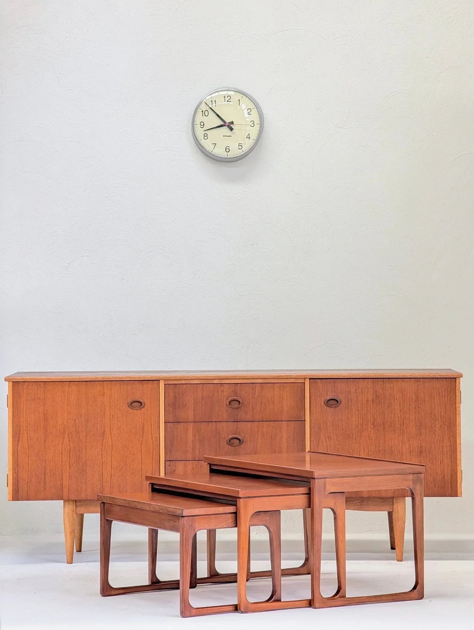 Mid-century teak nesting tables and sideboard against a white wall with clock