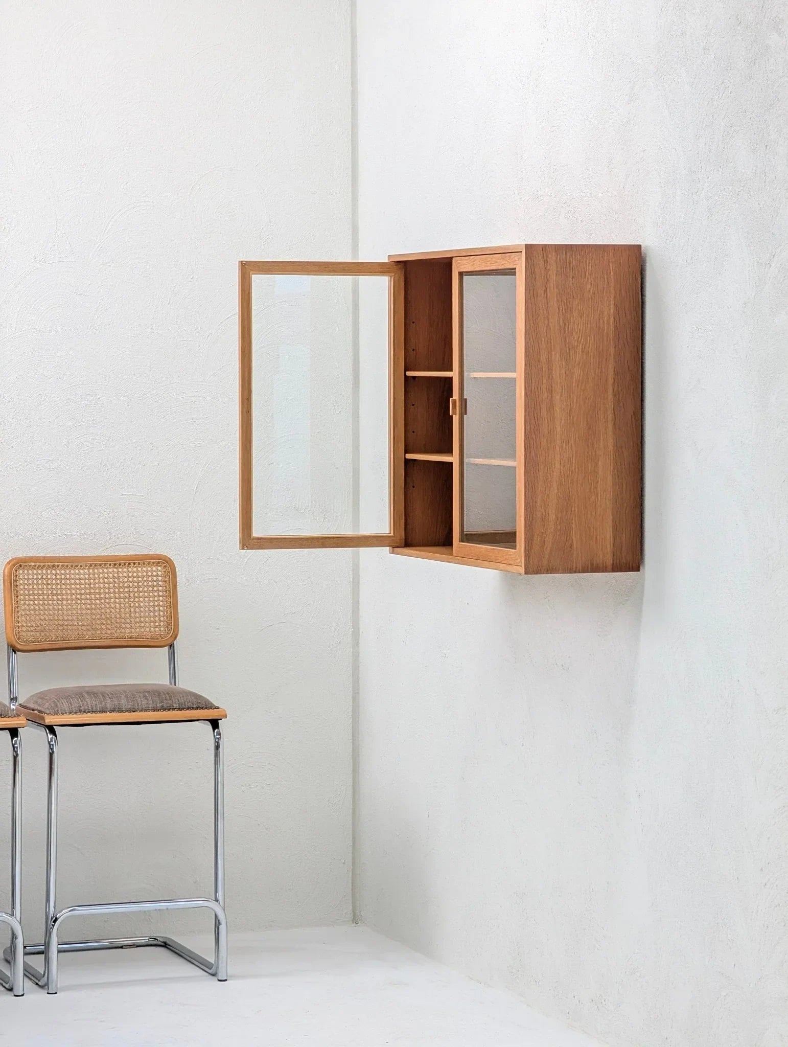 Mid-century oak and glass display cabinet mounted on a white wall, next to a woven chair