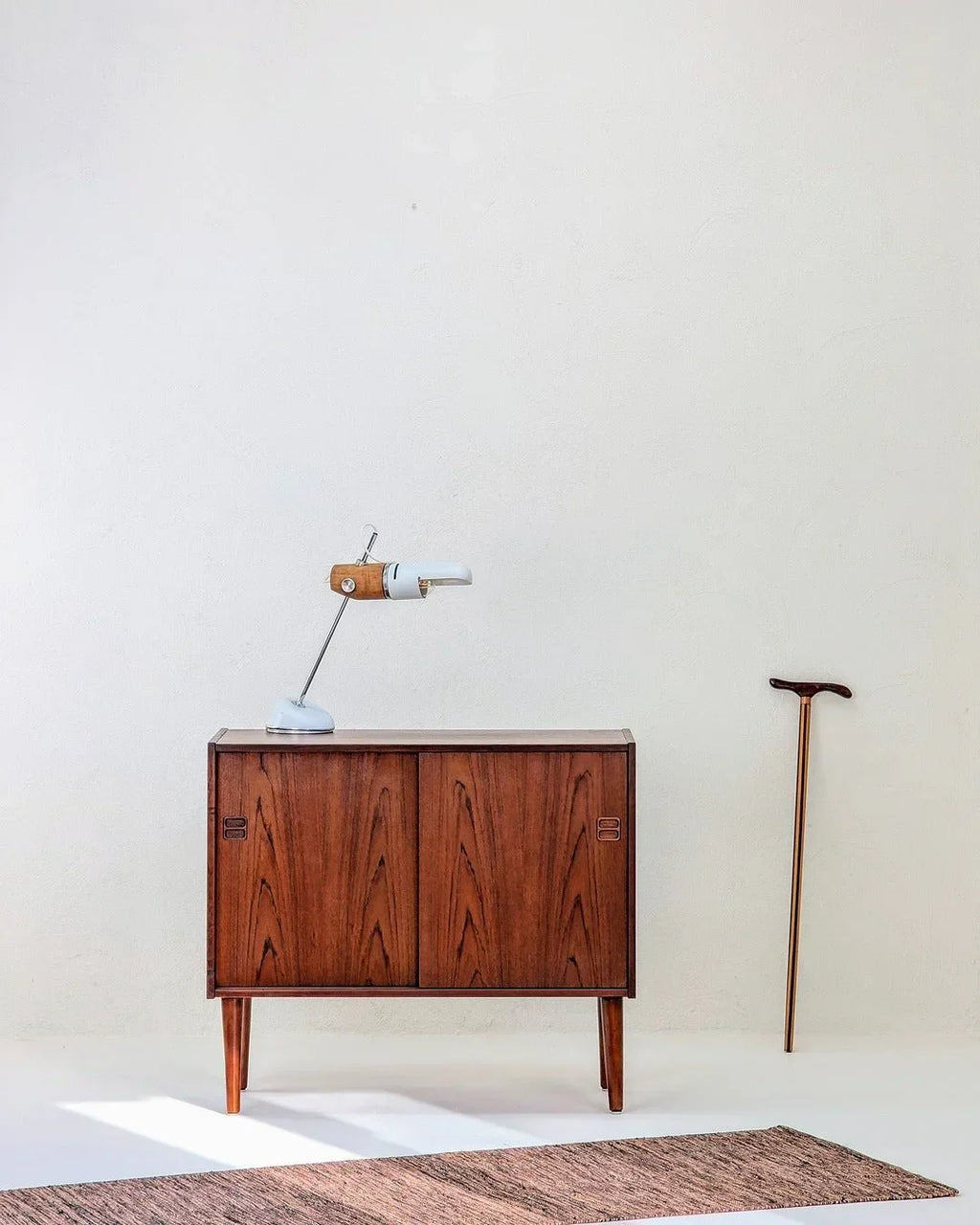 Mid-century Danish teak cabinet with lamp and walking cane against white wall