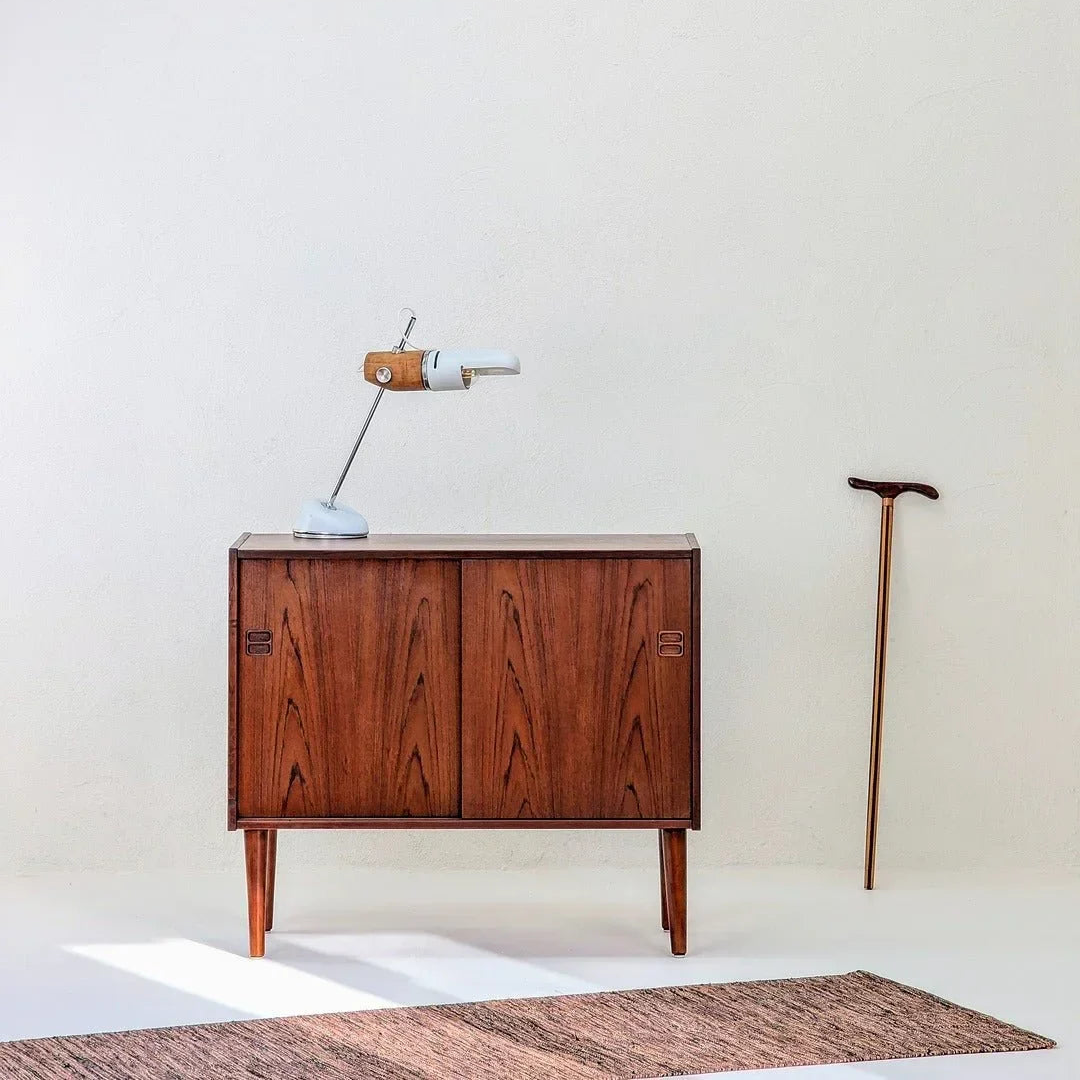 Mid-century Danish teak cabinet with sliding doors, vintage desk lamp on top and wooden walking cane against white wall