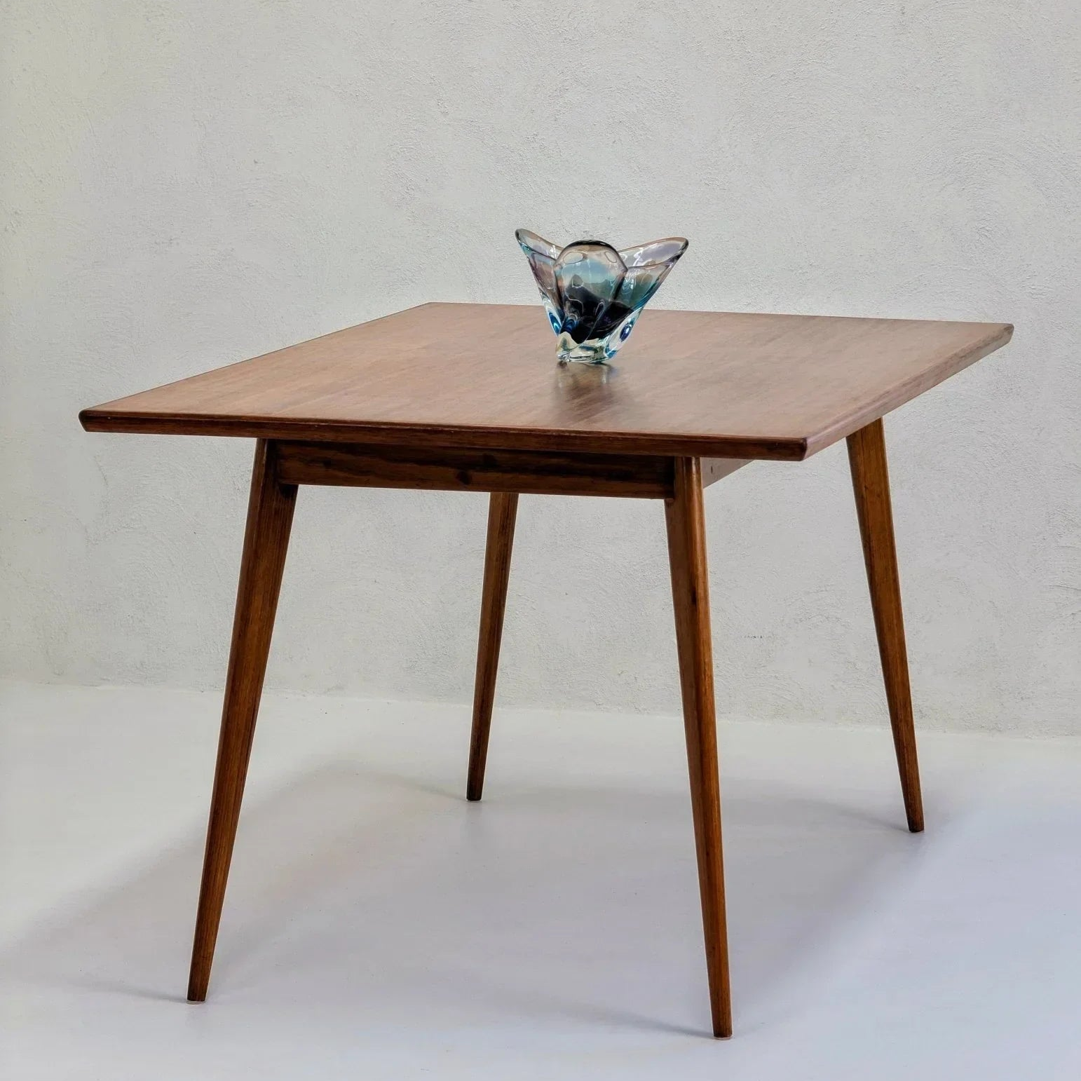 Mid-century modern square dining table in golden teak wood with tapered legs and a decorative glass centerpiece
