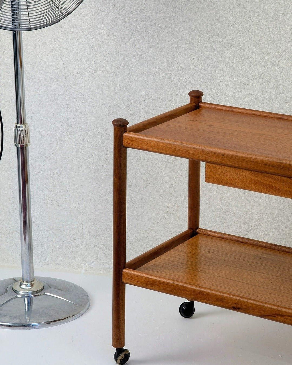Mid-century teak bar cart with wheels beside chrome floor fan against white wall