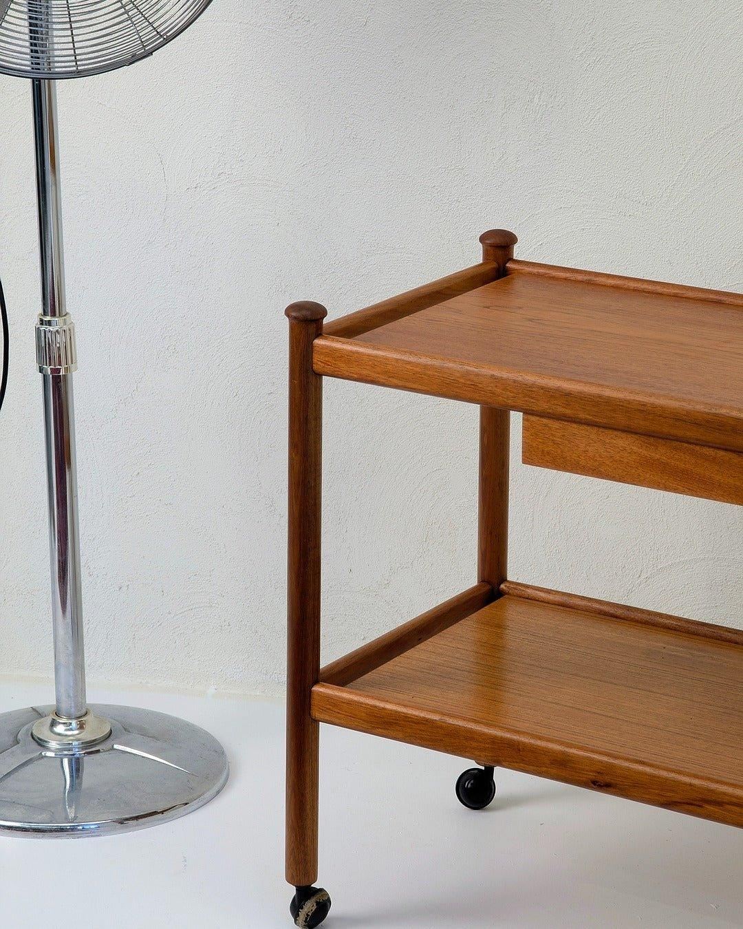 Mid-century teak bar cart with wheels beside chrome floor fan against white wall