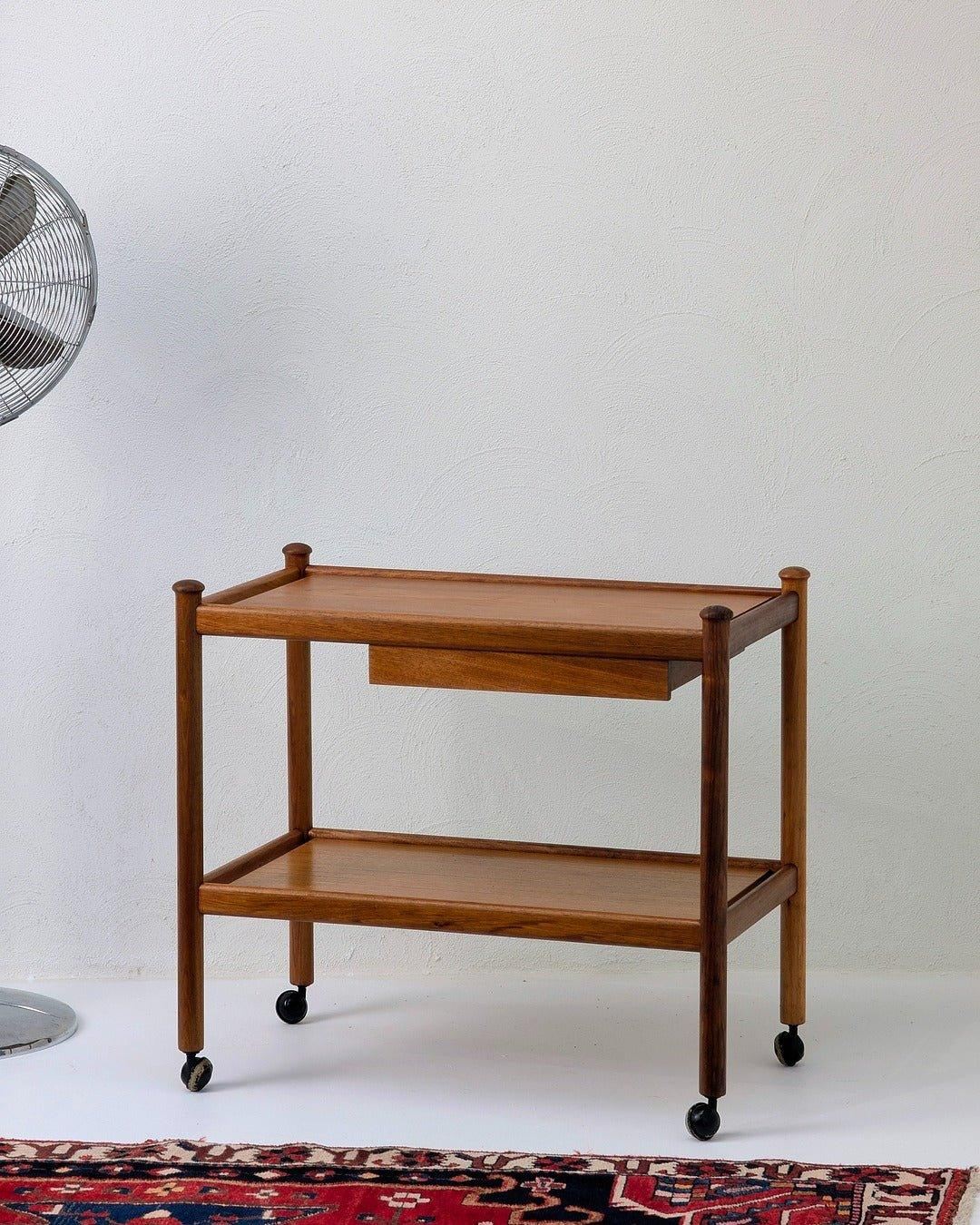 Mid-century teak bar cart with two shelves and wheels on a patterned rug