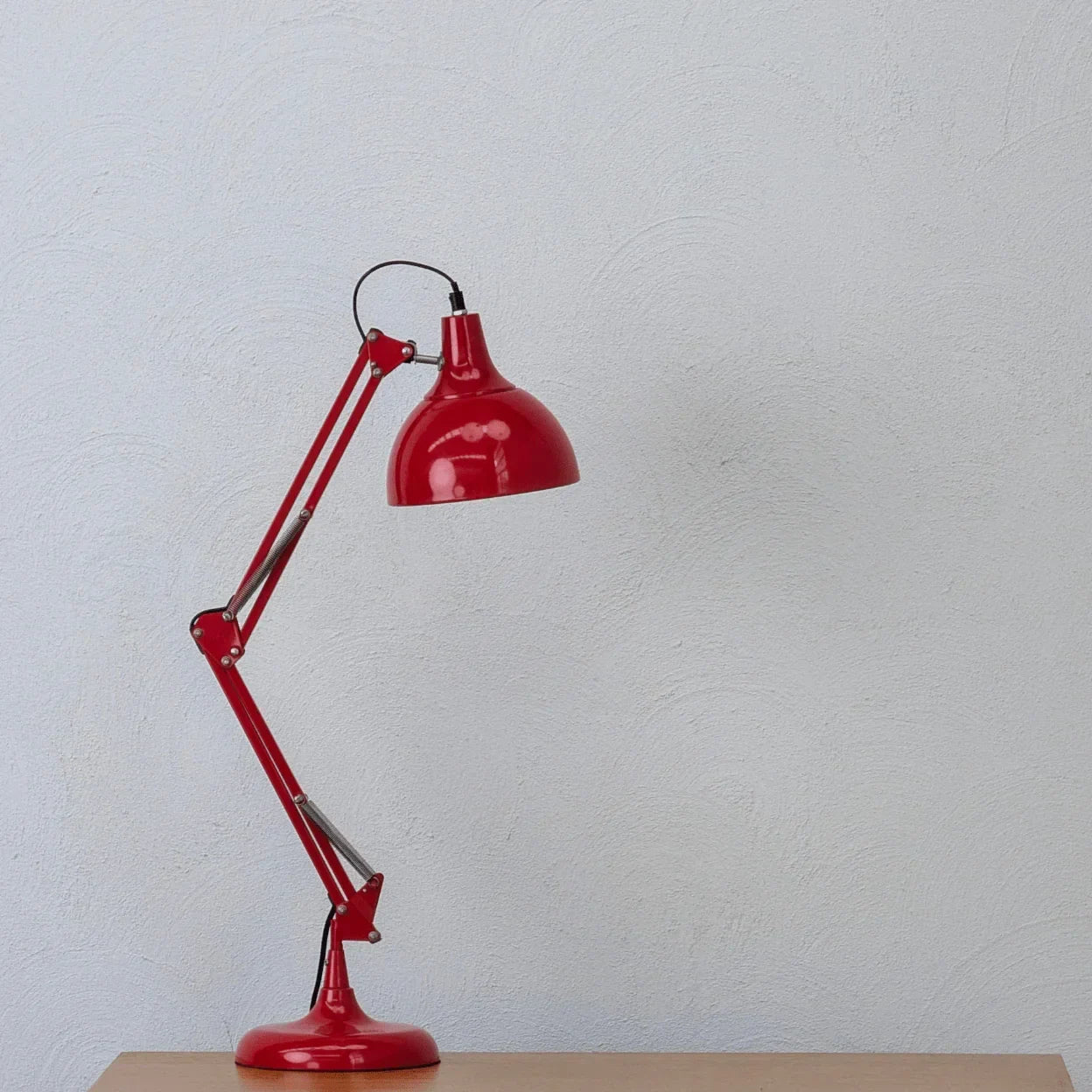 Red retro articulated desk lamp on a light wood table against a plain white wall