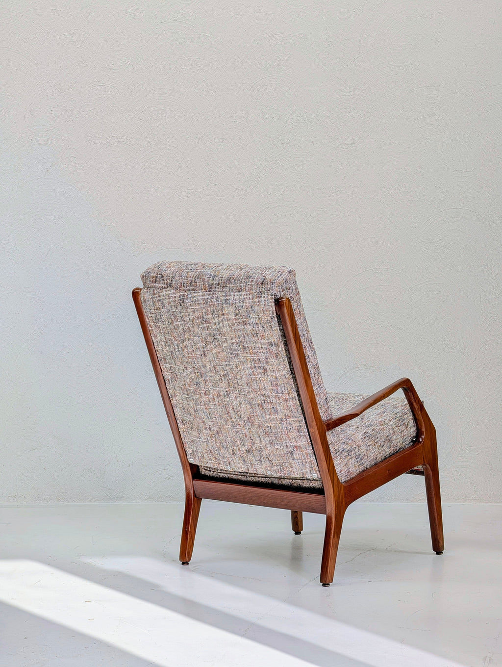 A mid-century modern accent chair with a warm brown wooden frame and textured tweed upholstery in beige, pink, and blue, shown against a minimalist off-white background.