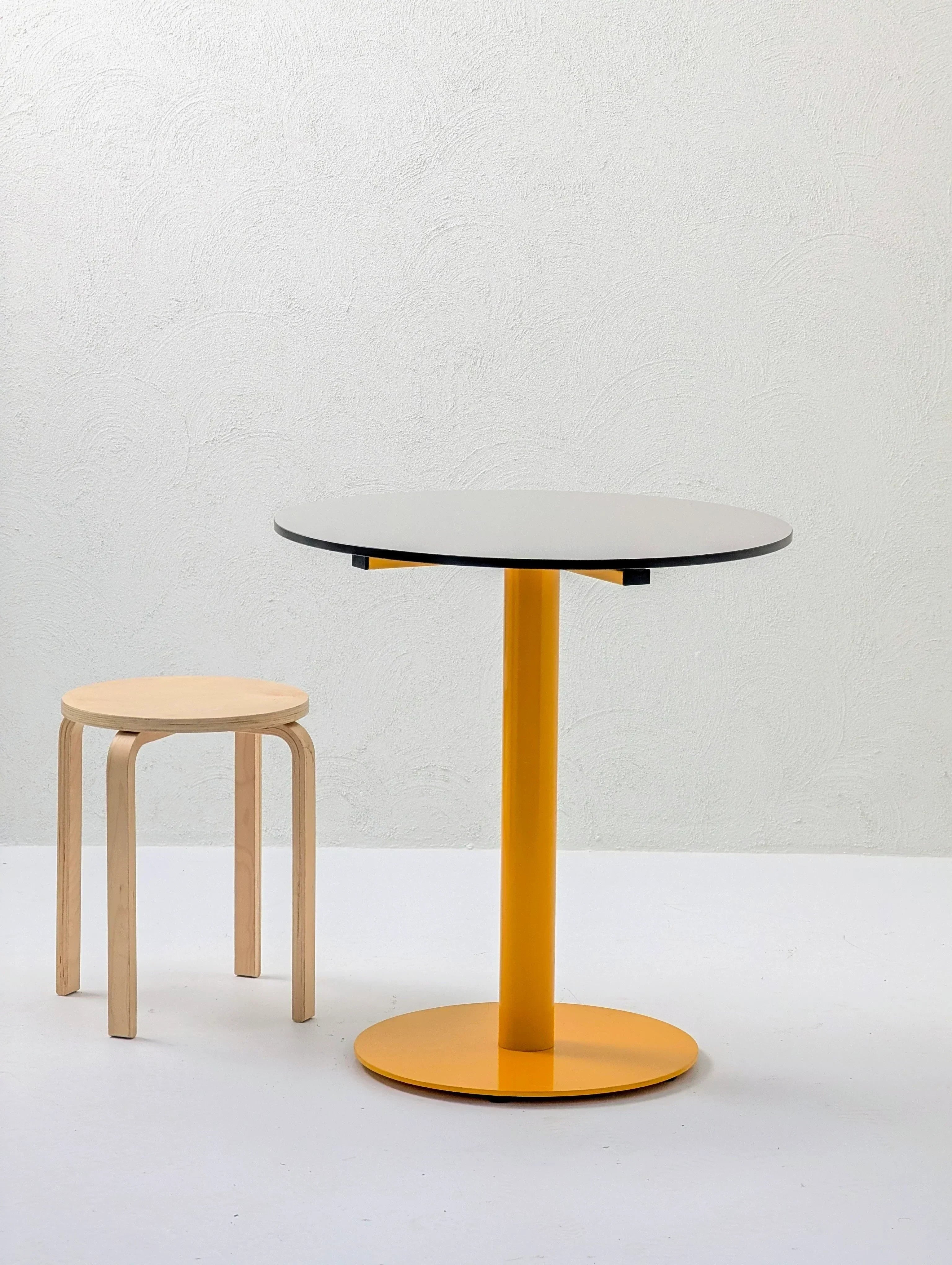 Minimalist scene with a round yellow pedestal table and a natural wood stool on a white floor