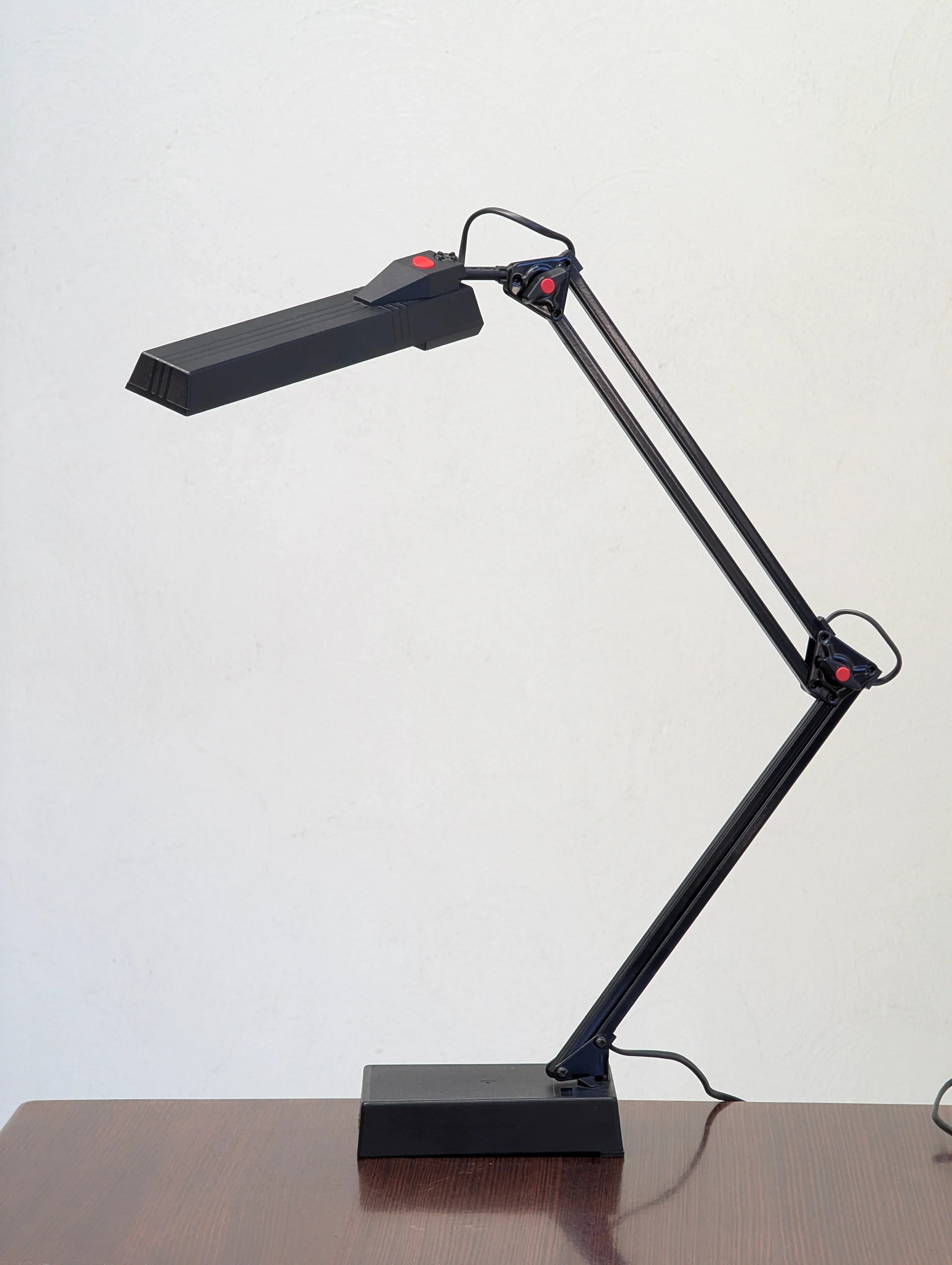 Black adjustable desk lamp with red buttons on a wooden table against a white wall