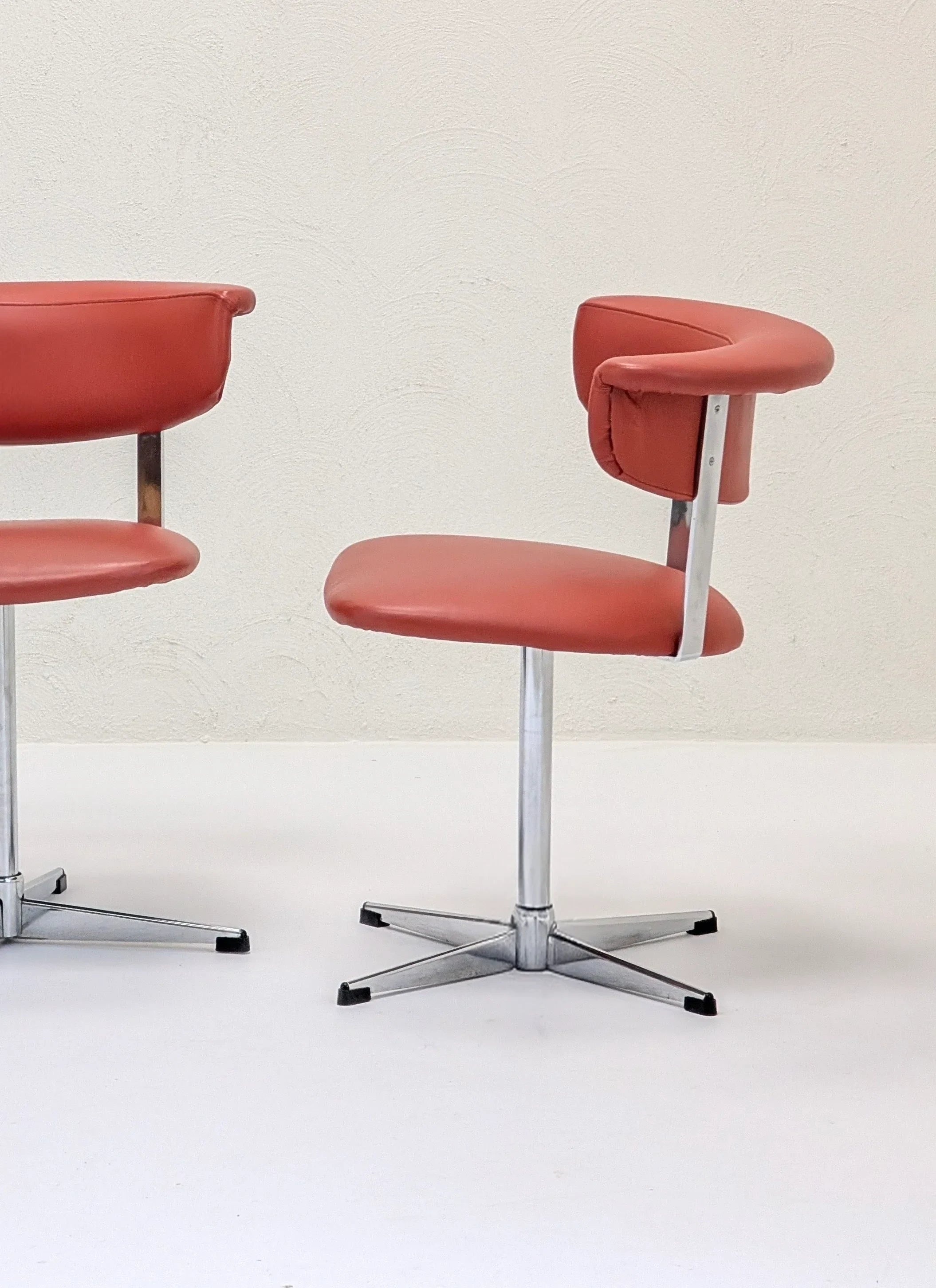 Two mid-century modern office chairs with orange upholstery and chrome swivel bases on white floor