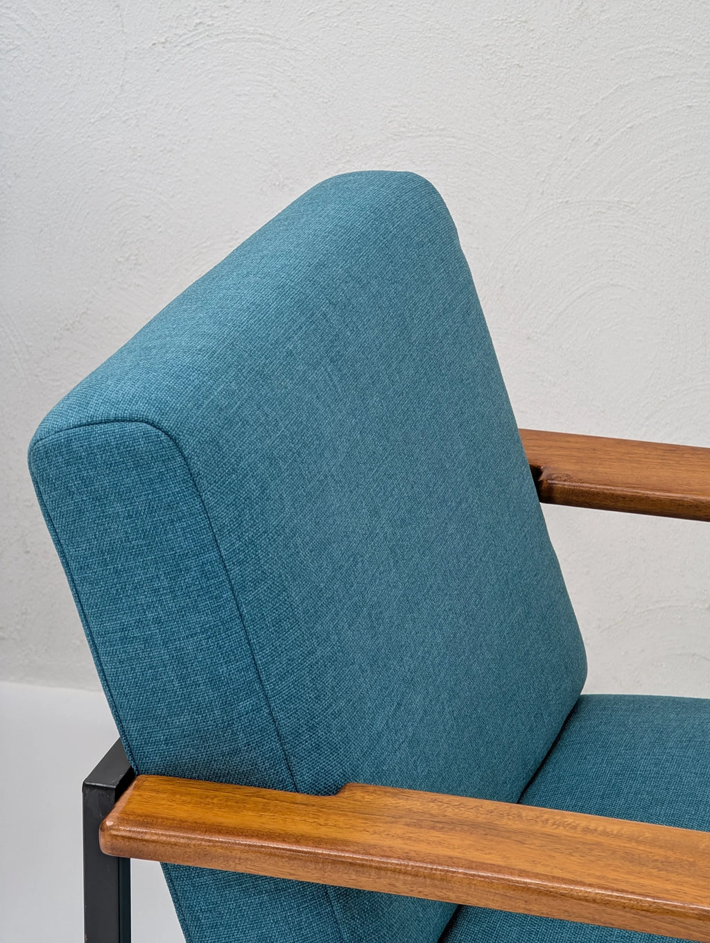 Mid-century modern chair with teal upholstery and wooden armrests against a textured wall