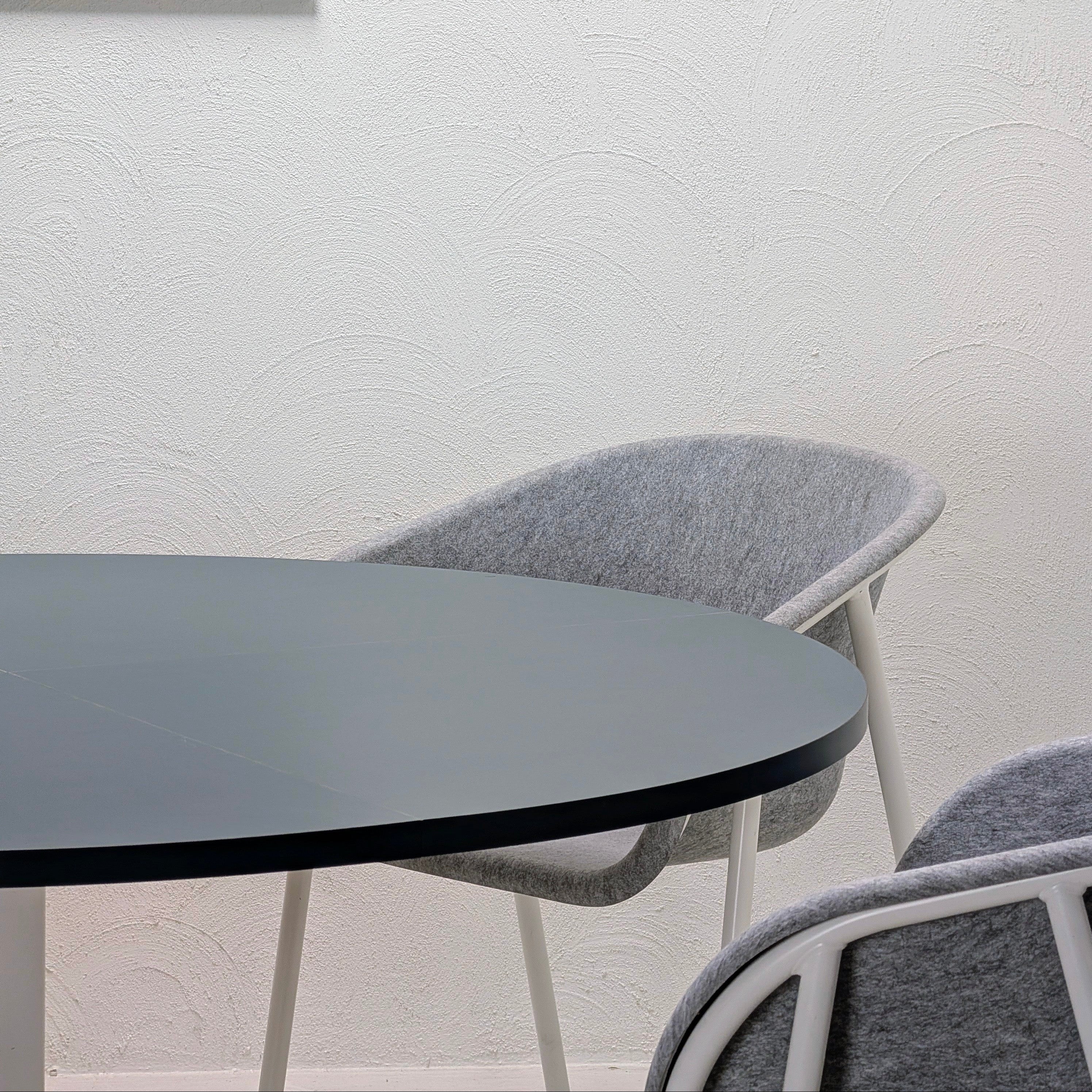 A modern dining setup featuring a round black table and two gray upholstered chairs with white frames, set against a textured off-white wall.