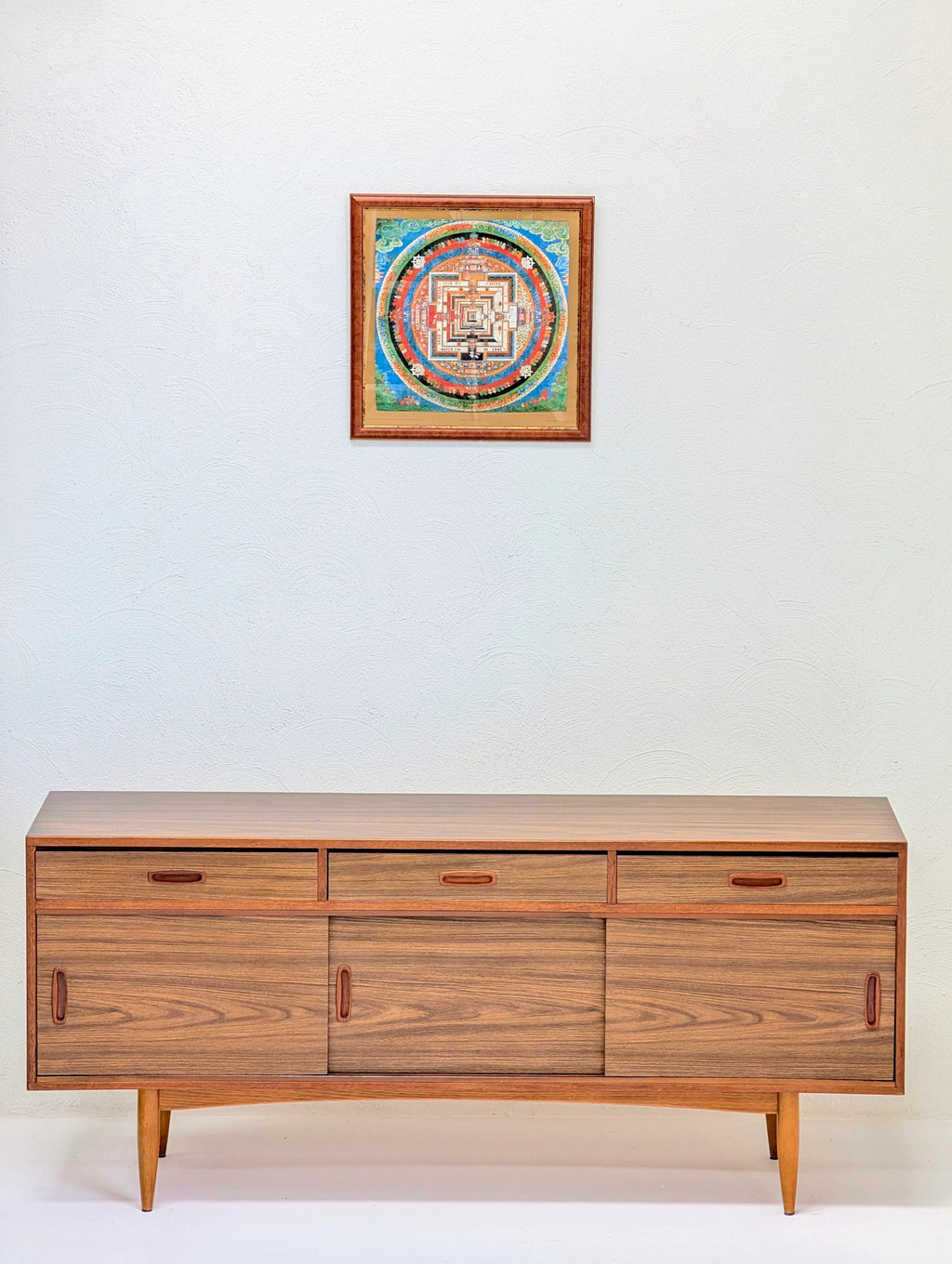 Mid-century modern wooden sideboard with sliding doors and three drawers under framed circular mandala artwork