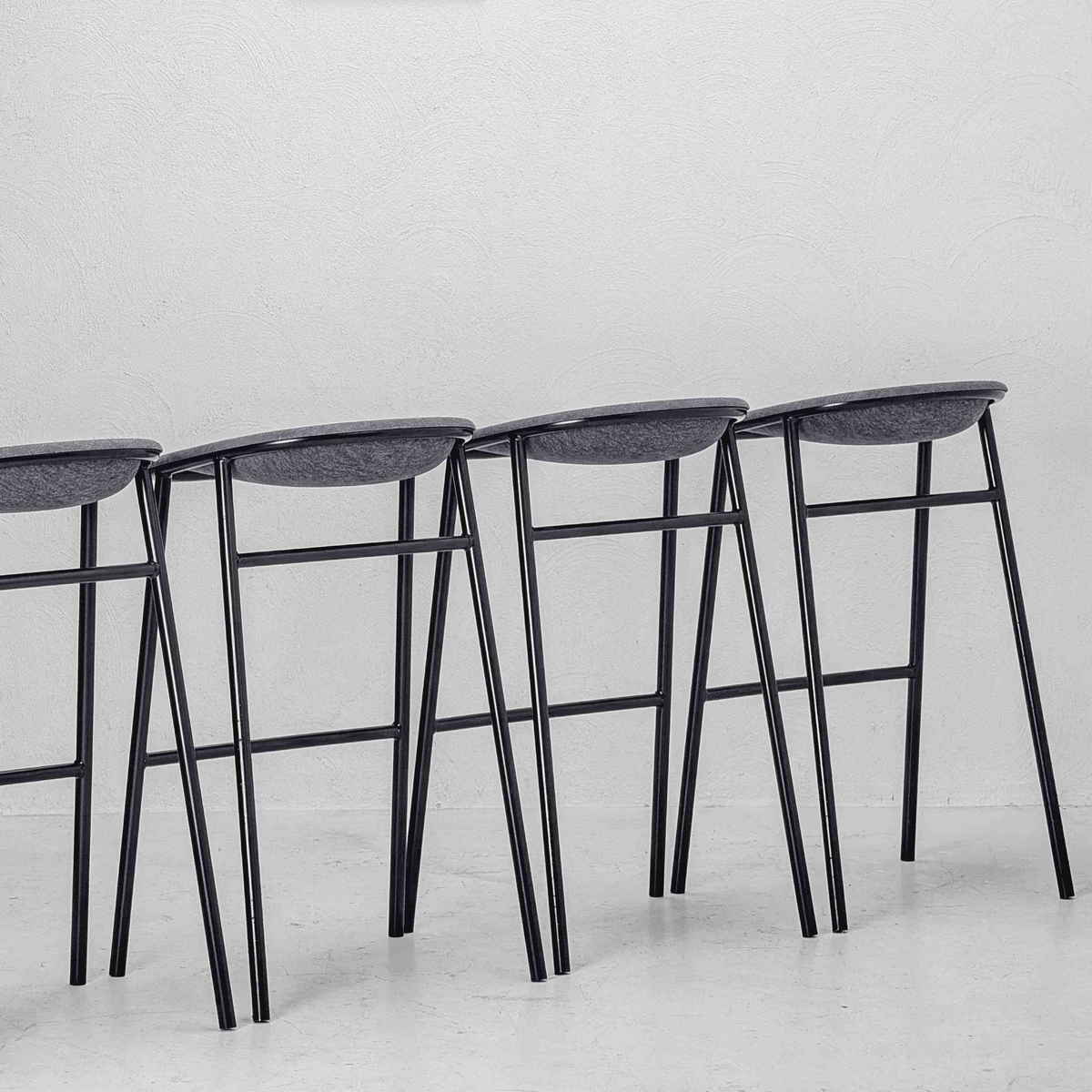 Four modern bar stools with dark gray oval seats and black metal legs, arranged in a row against a white textured wall.

