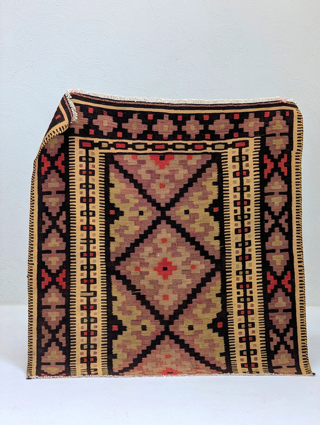 Traditional geometric woven rug with black, red, beige, and brown patterns displayed against a white wall