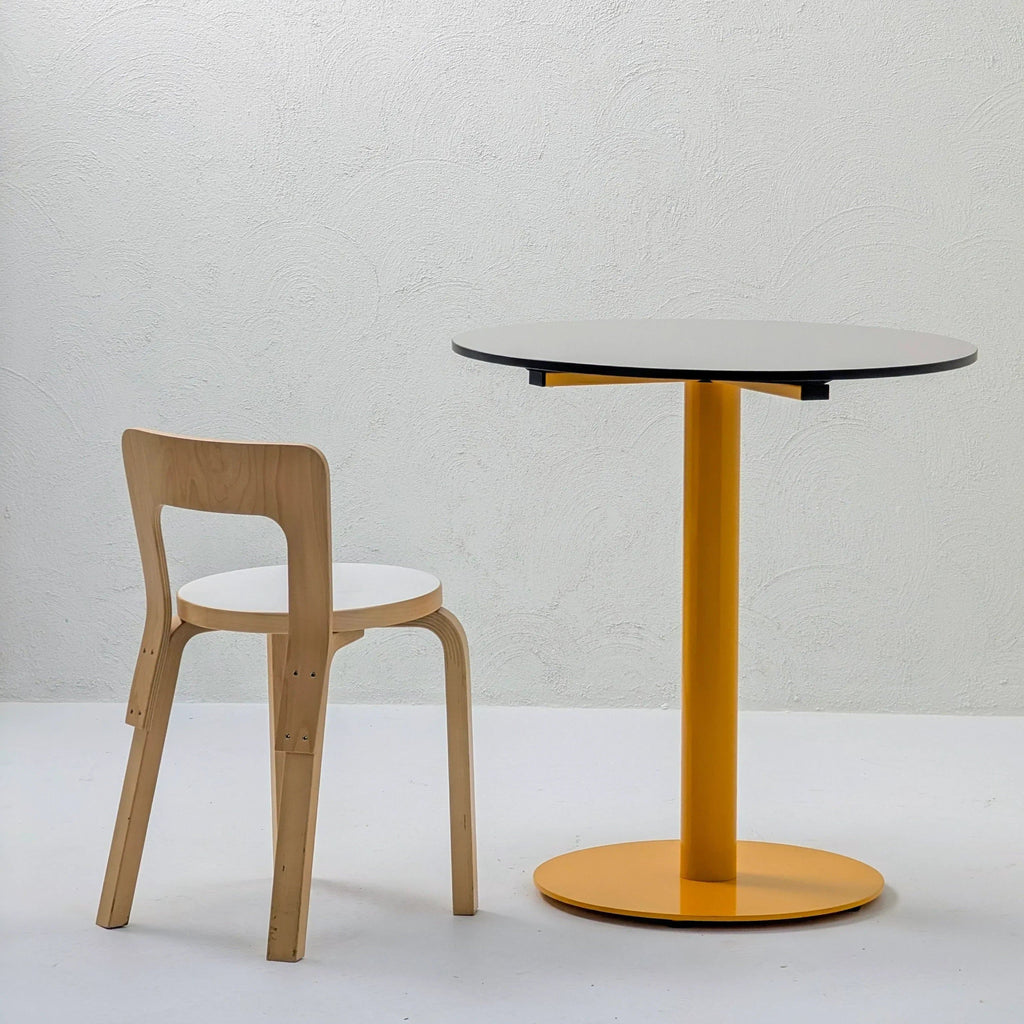 Minimalist wooden chair and round table with yellow base against a textured white wall