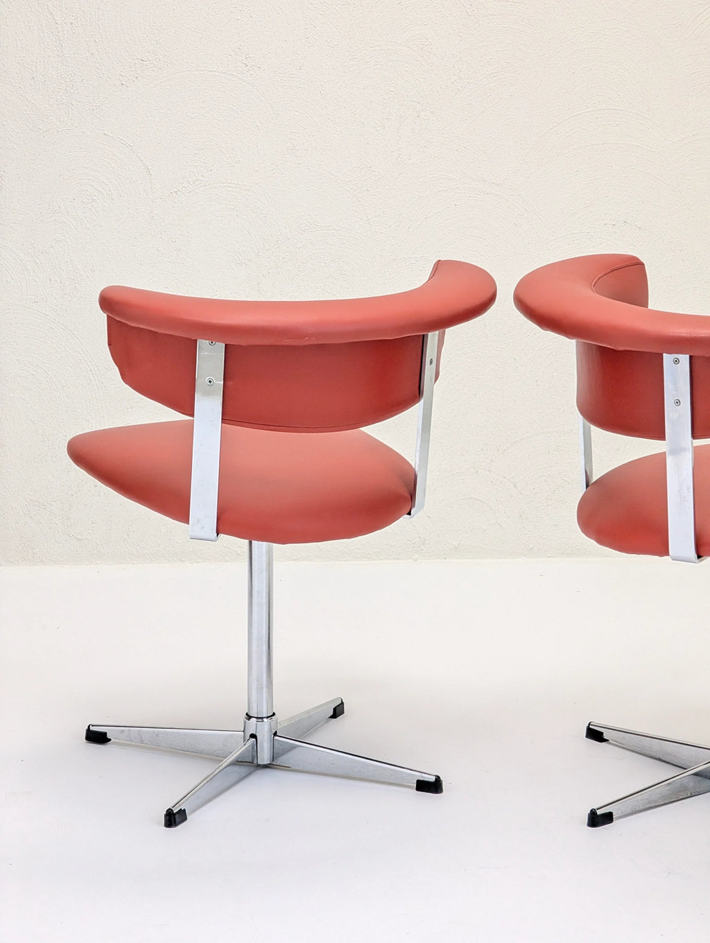 Two modern orange swivel chairs with chrome legs on a white floor and textured beige wall