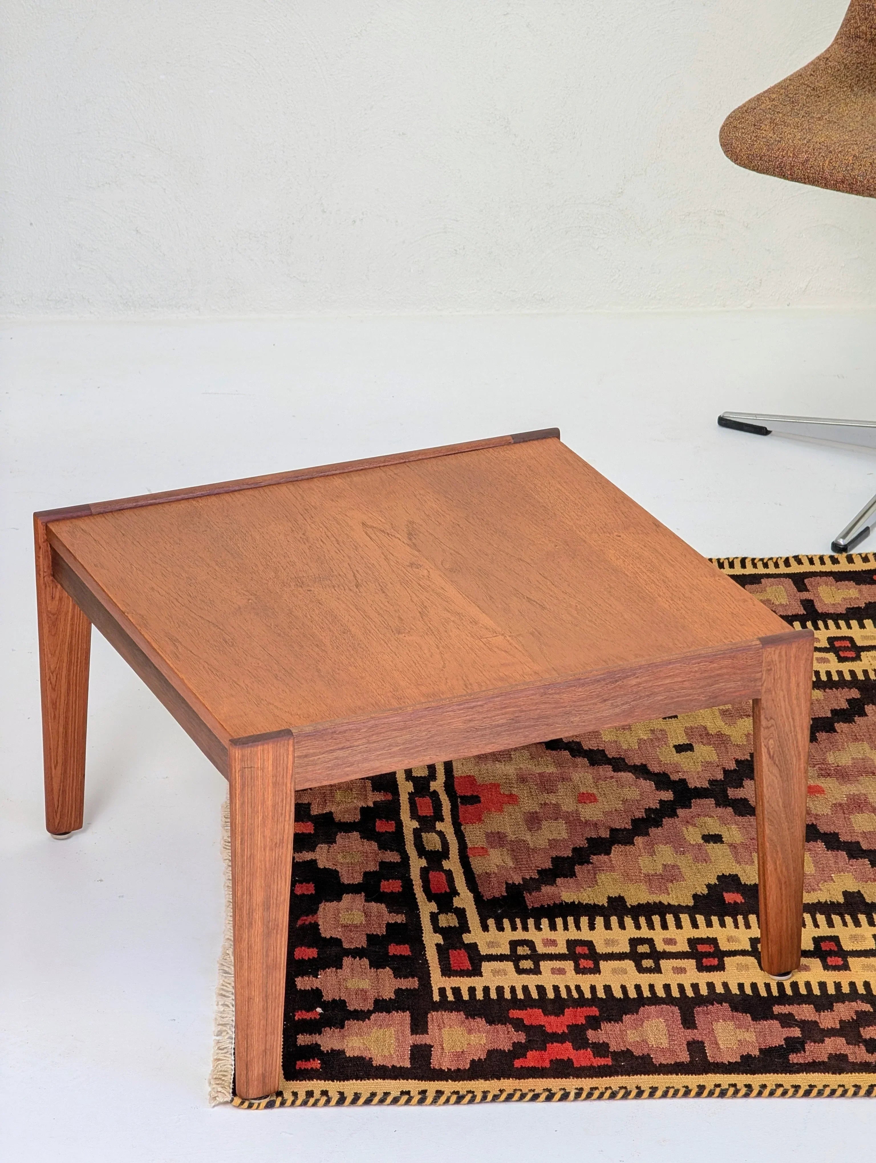 Square wooden coffee table on a patterned woven rug with a mid-century modern chair nearby