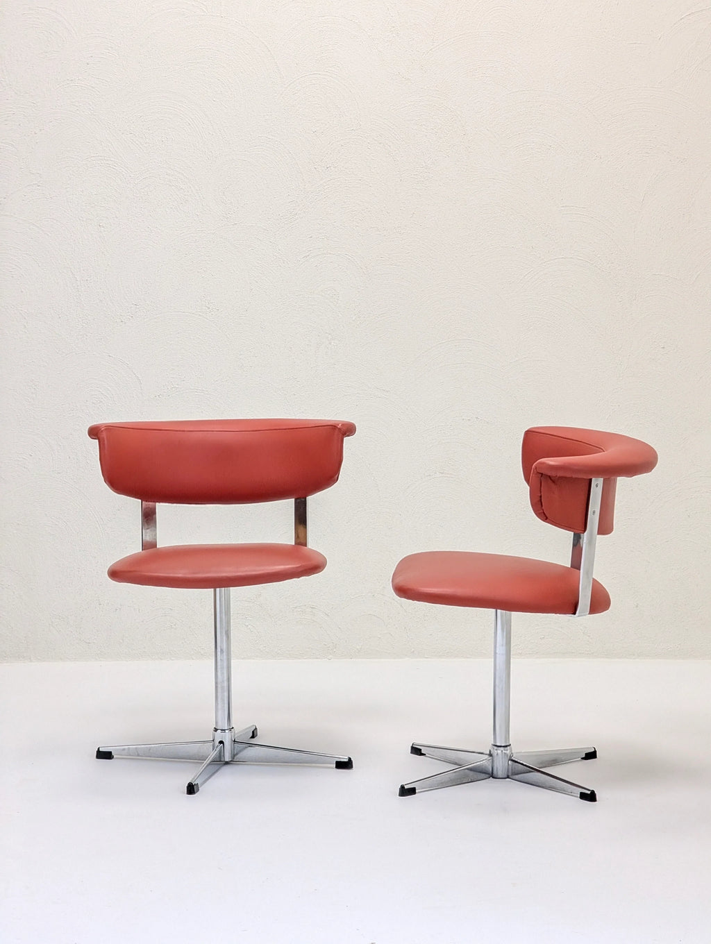 Two modern retro-style swivel chairs with orange cushions and chrome bases on white floor