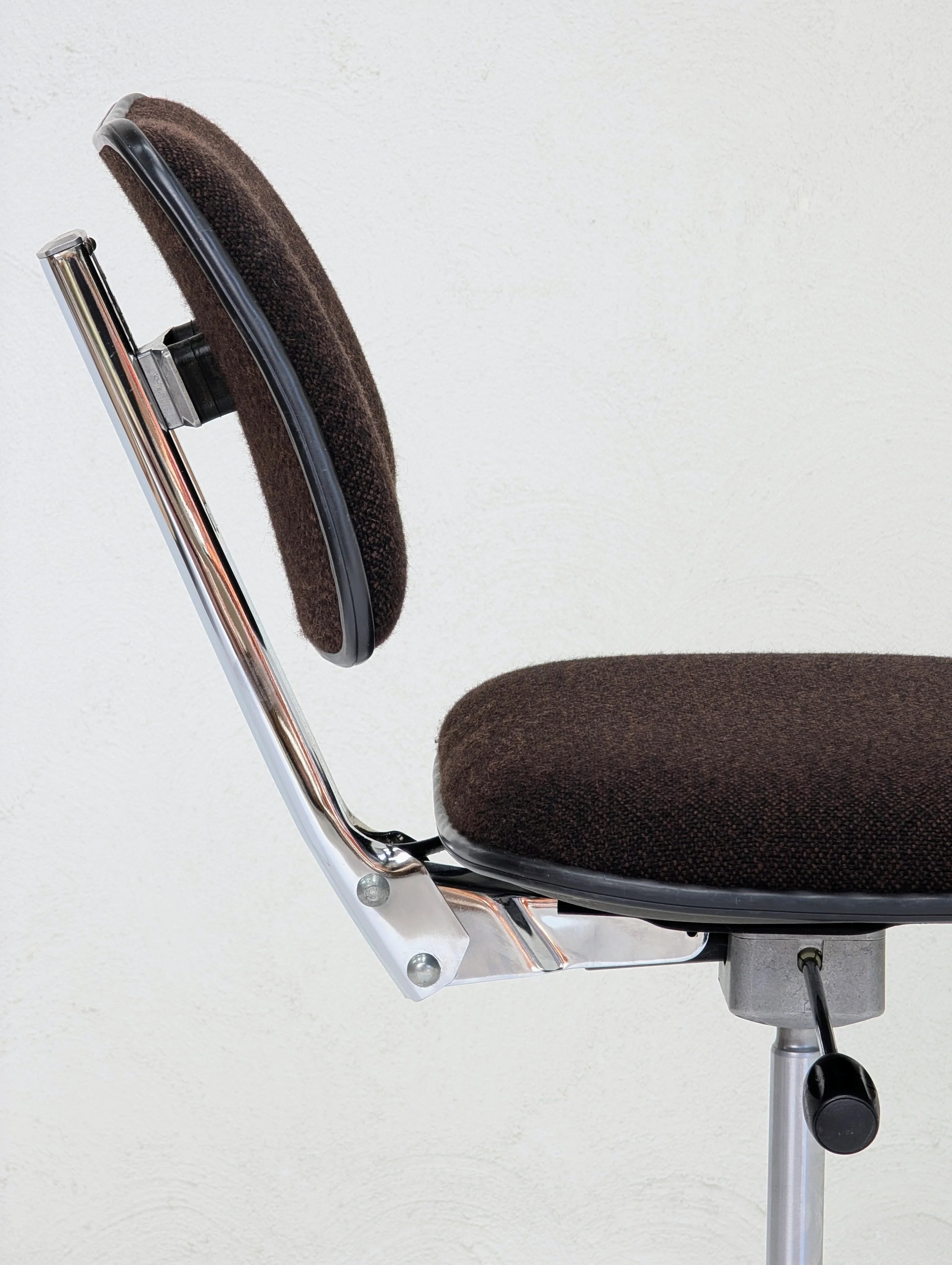 Side view of a brown fabric office chair with chrome frame and adjustable lever against white wall