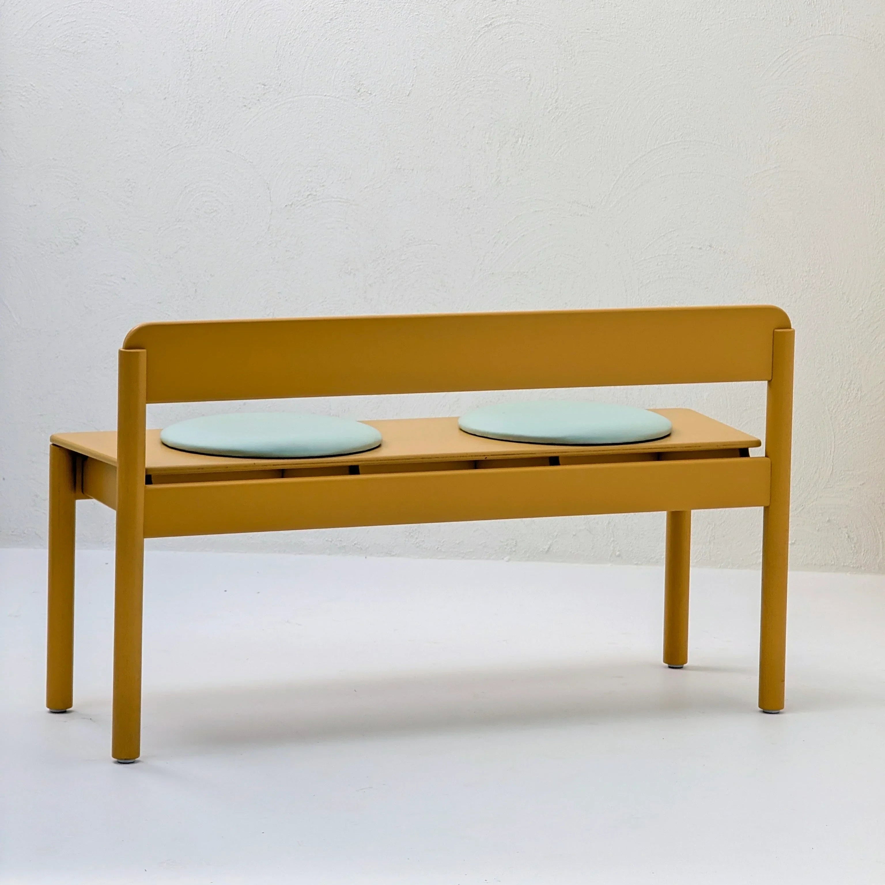 Modern yellow bench with backrest and two round mint seat cushions against white wall