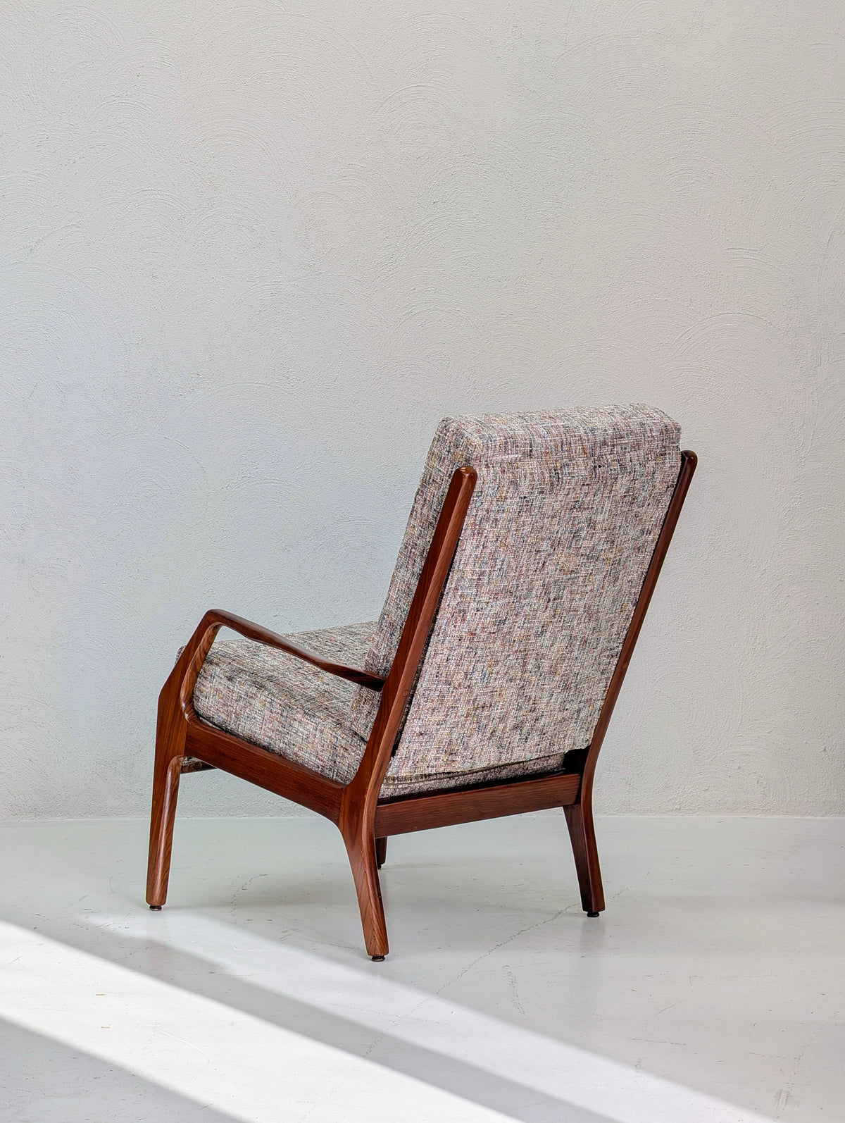 A mid-century modern accent chair with a warm brown wooden frame and textured tweed upholstery in beige, gray, and muted red tones, shown against a minimalist off-white wall and floor.