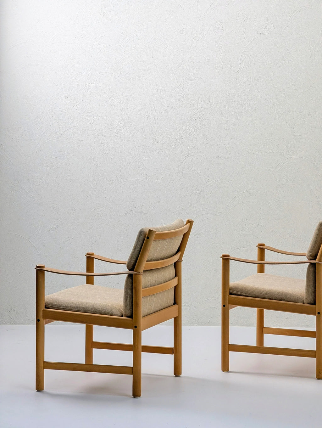 Two modern wooden armchairs with beige cushions against a light textured wall