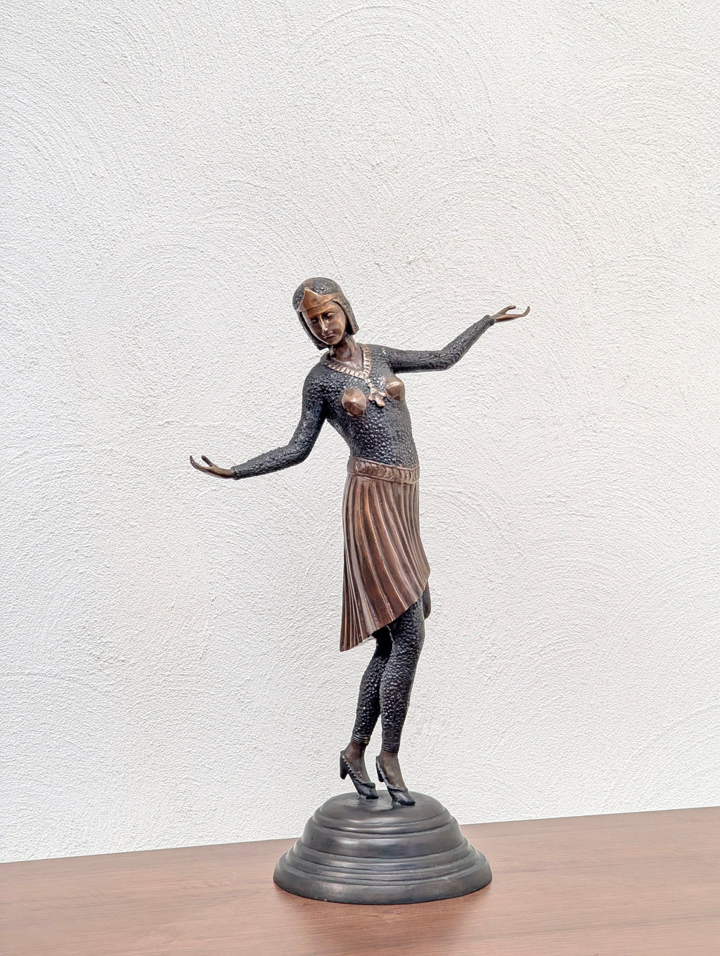 Bronze statue of a dancing woman with a pleated skirt on a round base, displayed on a wooden surface