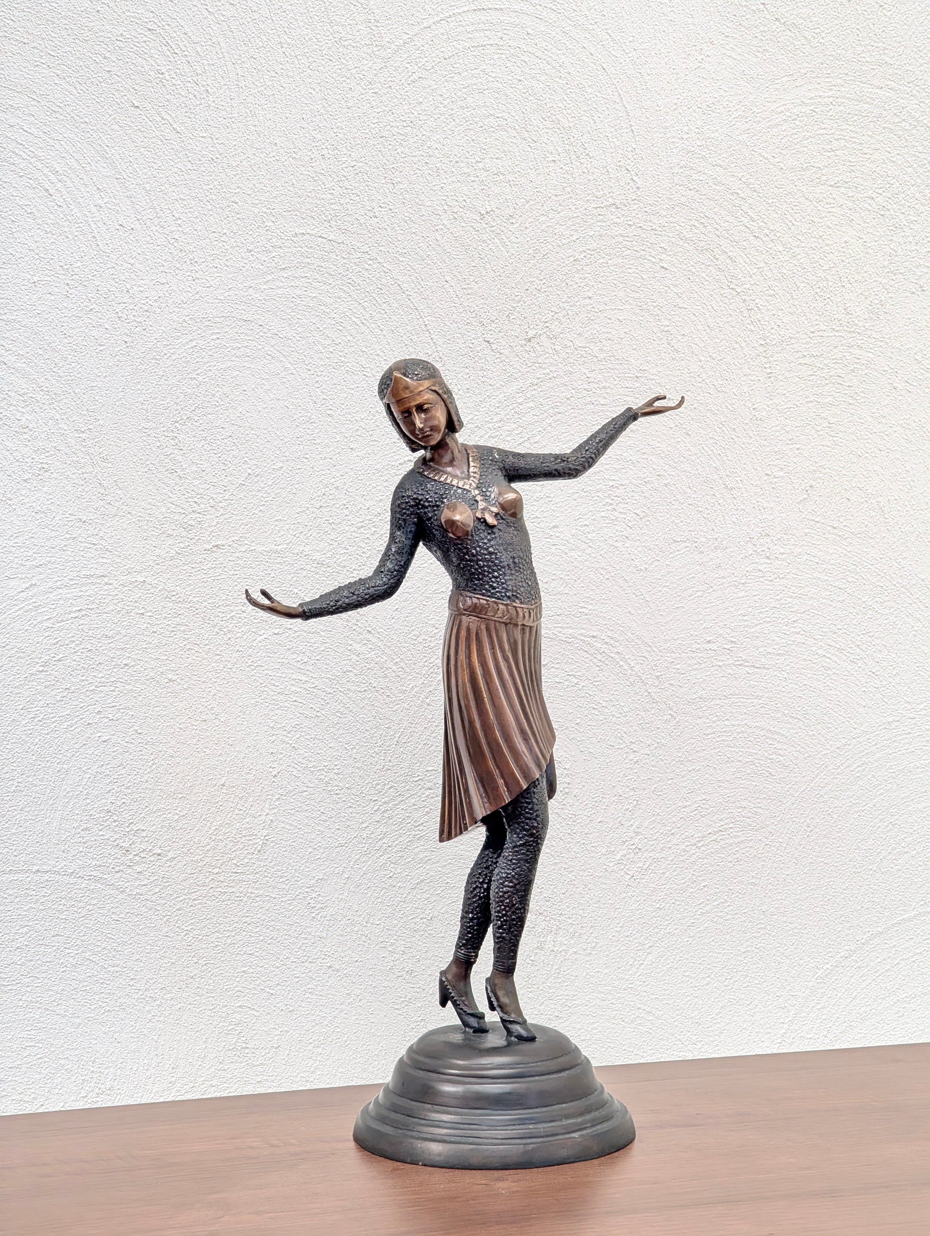 Bronze statue of a dancing woman with a pleated skirt on a round base, displayed on a wooden surface