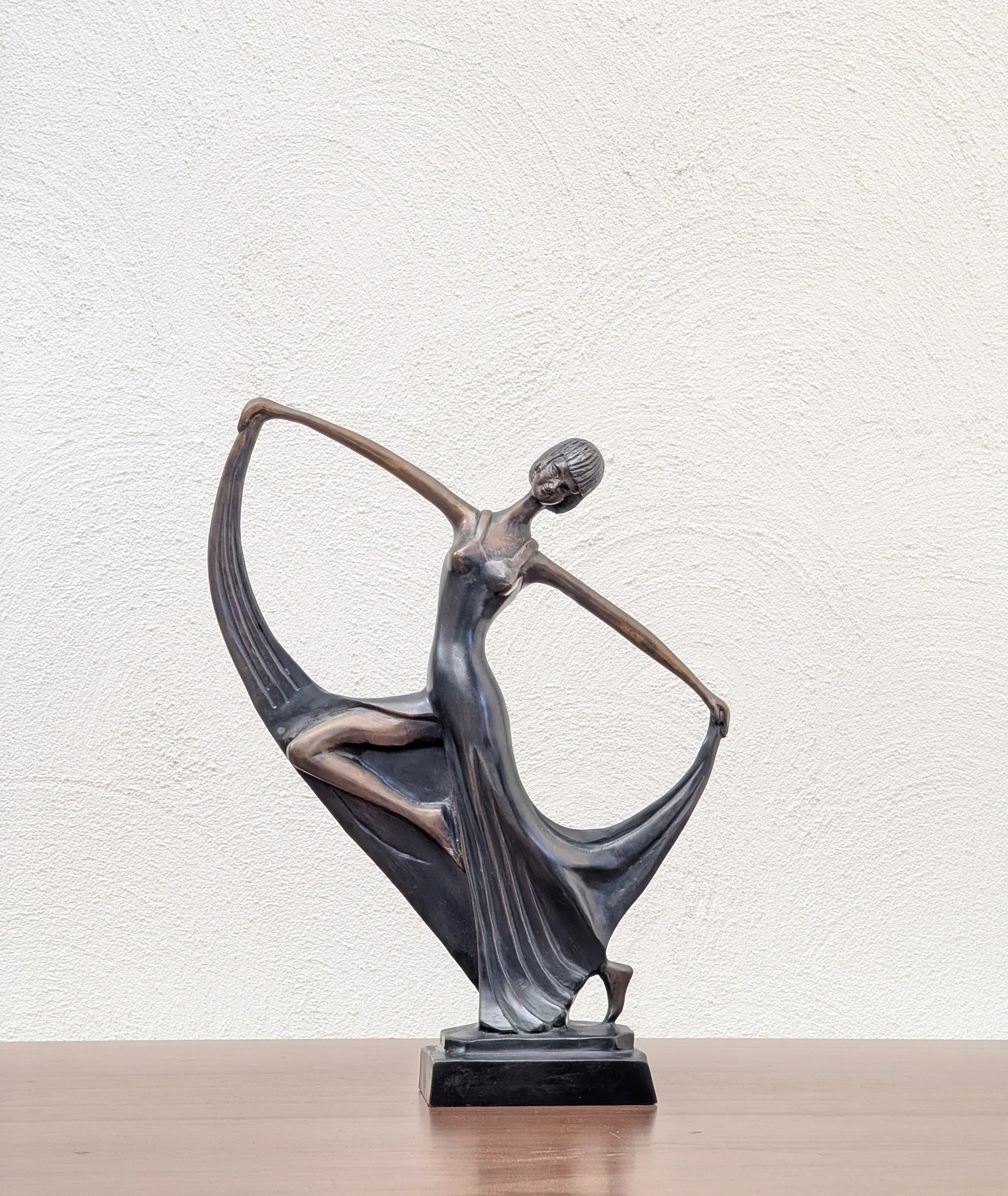 Bronze sculpture of a dancing woman in flowing dress on wooden surface with textured white wall
