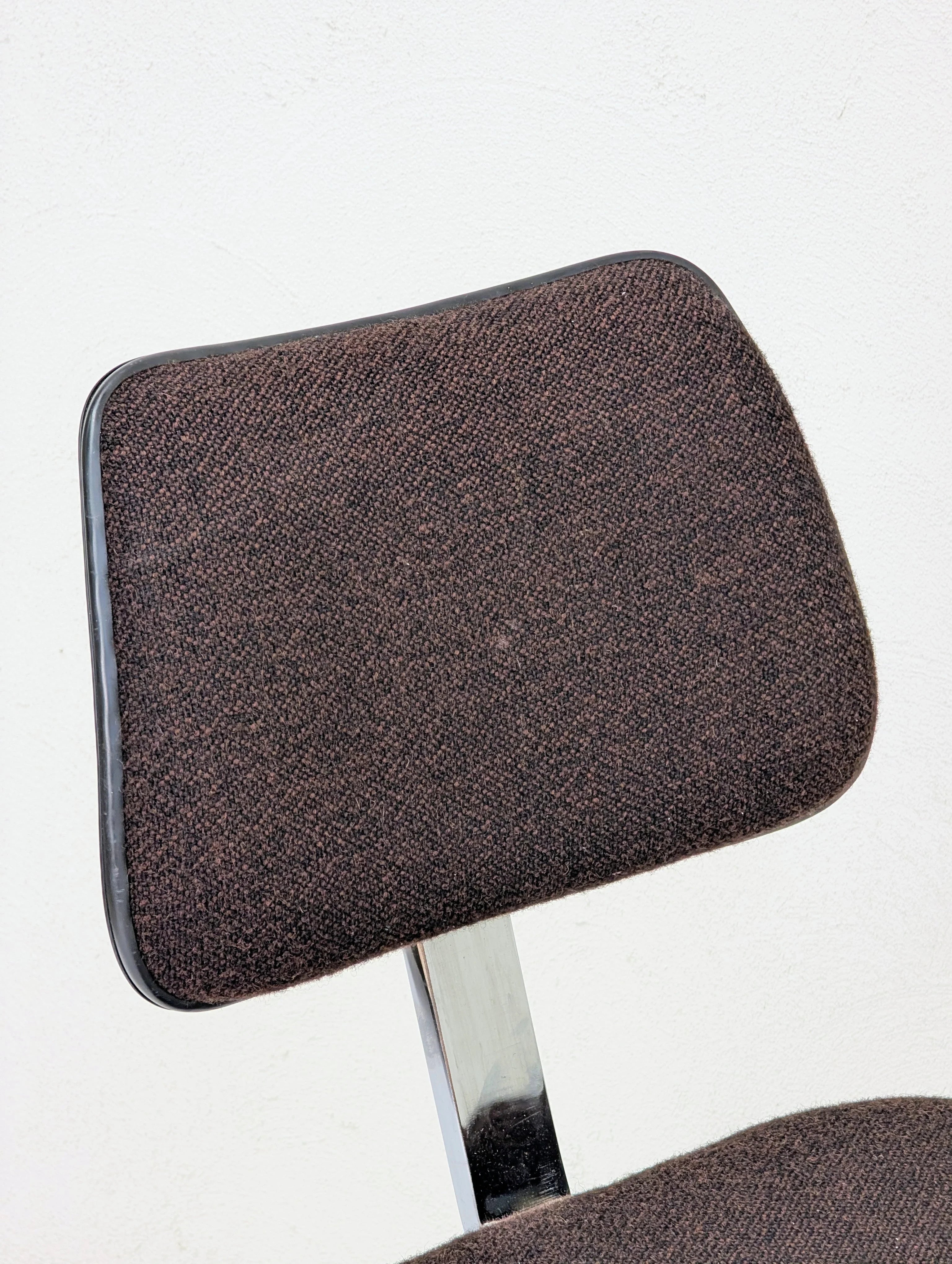 Close-up of a brown fabric office chair backrest with a chrome support against a white wall