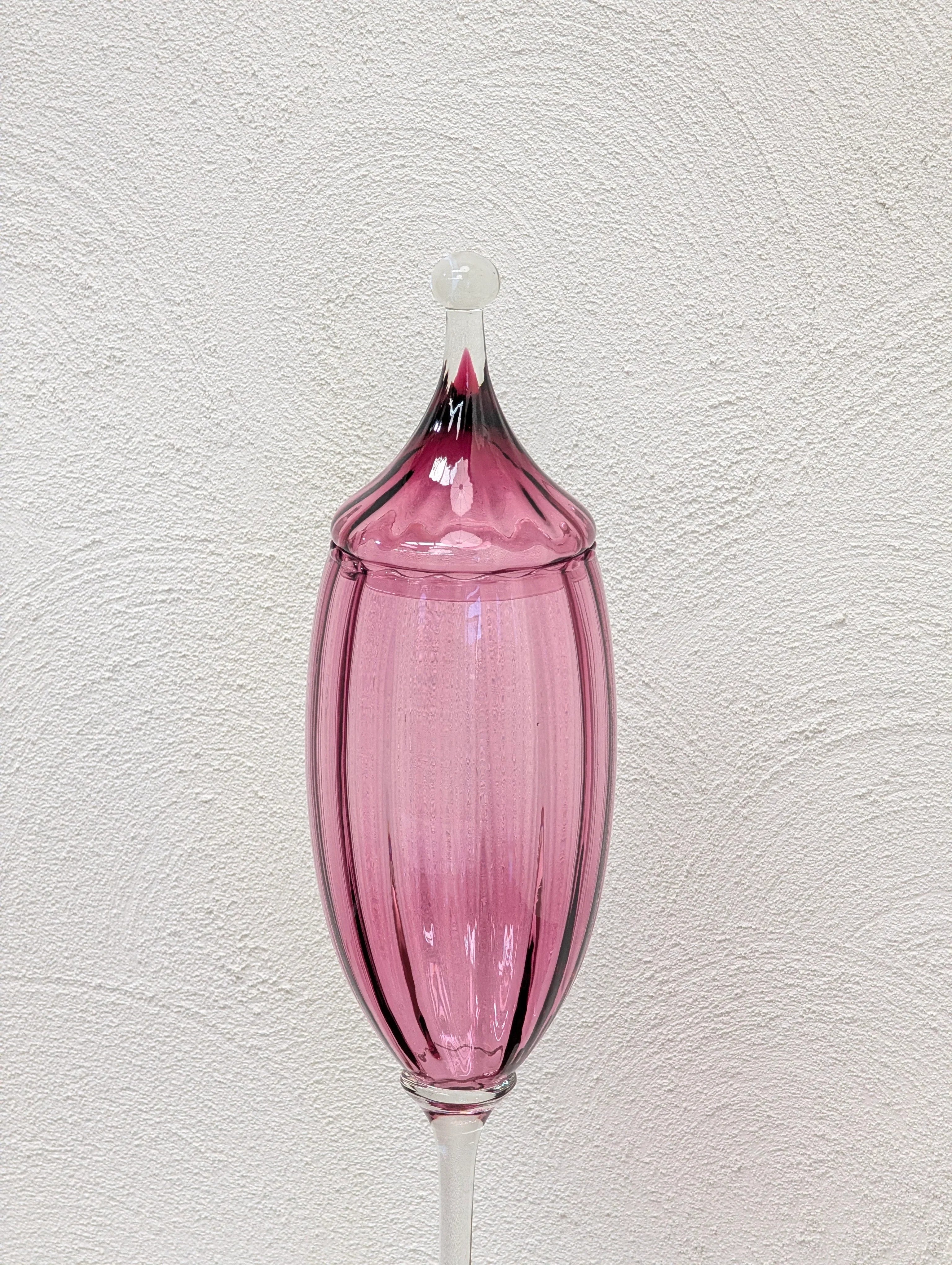 Elegant pink glass vase with pointed lid on textured white wall background