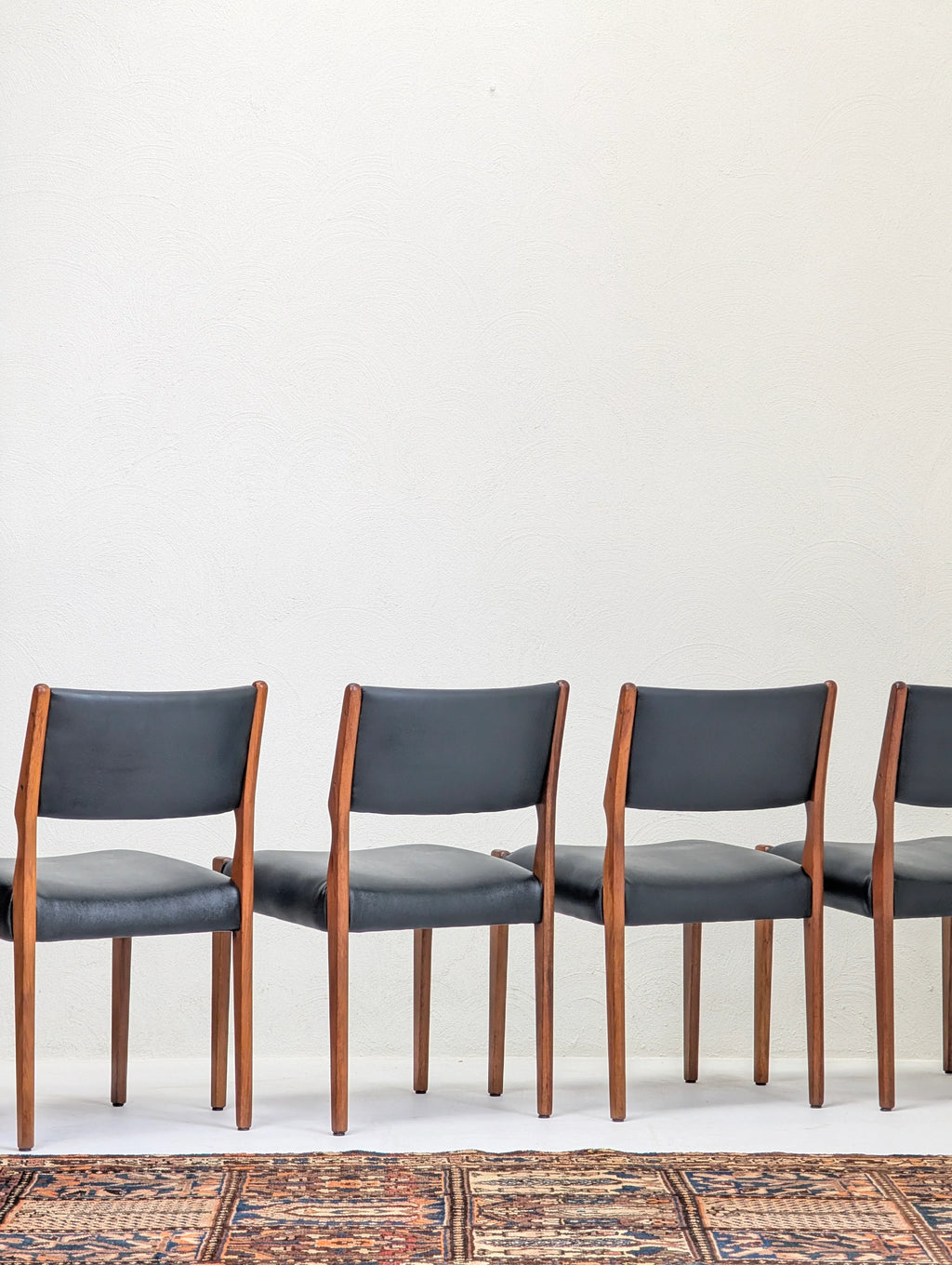 Four mid-century modern wooden chairs with black cushions lined up against a white textured wall on a patterned rug