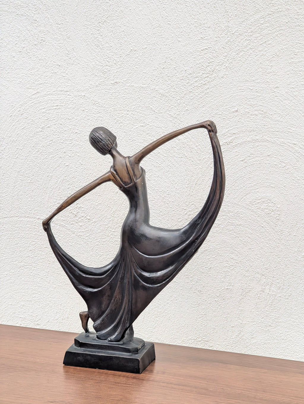 Bronze sculpture of a dancing woman with flowing dress on wooden surface against textured white wall